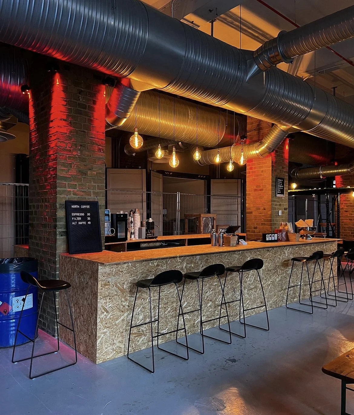 Modern coffee bar with copper textured counter, black chairs, exposed industrial ductwork, and warm Edison bulb lighting.
