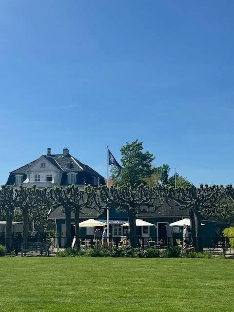 A large house with a steep roof and dormer windows in the background, a flagpole with a flag in front, trees, an outdoor patio with umbrellas and seating, and a well-maintained grassy lawn under a clear blue sky.