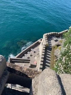 A scenic view of a coastal area with stairs leading down to the water, surrounded by greenery and rocky cliffs.
