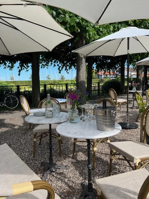 Outdoor patio dining area with white umbrellas, marble tables, flowers in vases, and chairs surrounded by trees and greenery.
