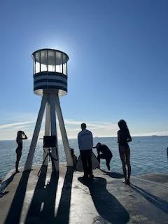 People standing and sitting near a lighthouse on a waterfront, with the ocean and clear sky in the background.