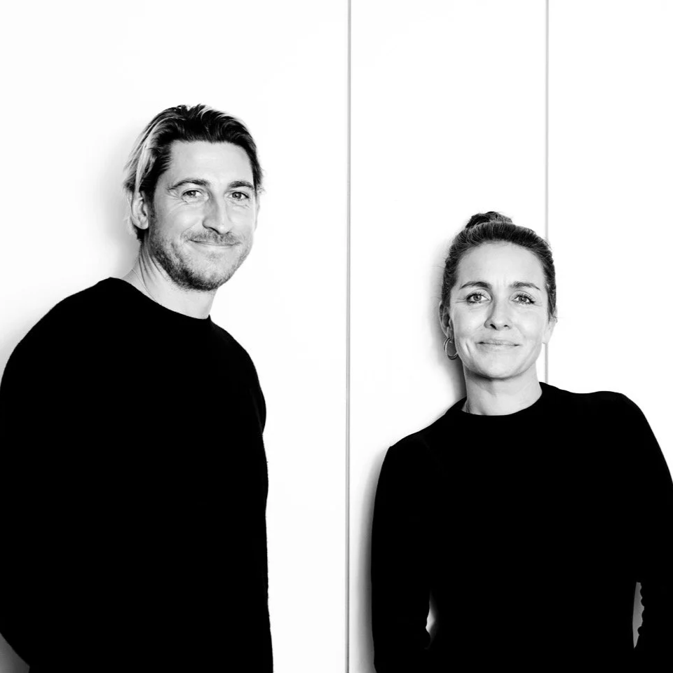 Black and white photo of a man and woman leaning against a plain wall, smiling at the camera.
