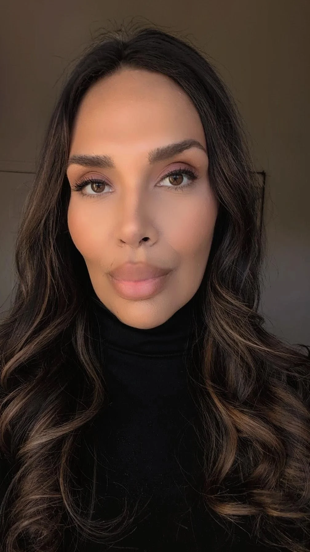 A Latina with long dark hair styled in loose waves, wearing a black turtleneck, looking directly at the camera with a neutral expression.