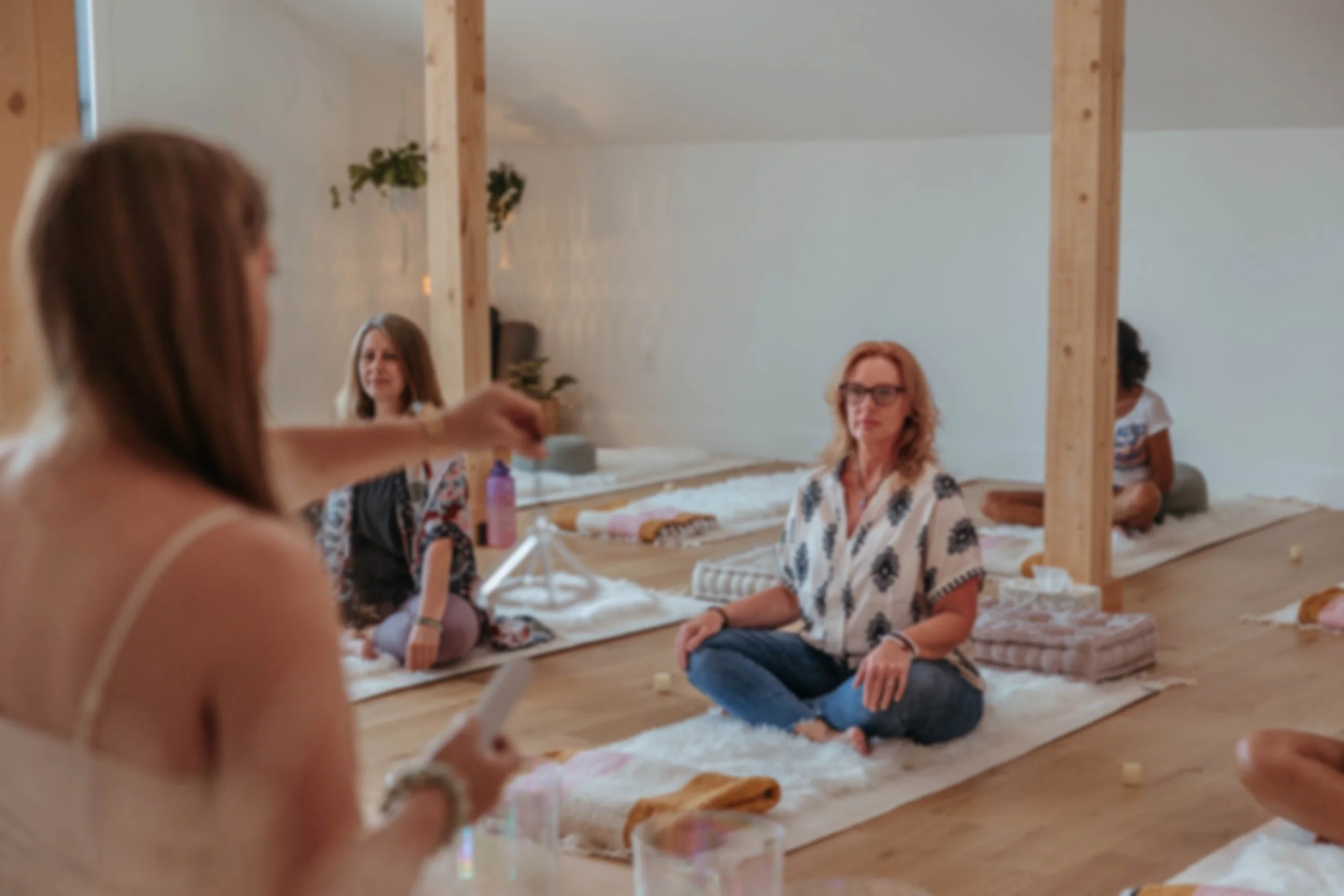 Women participating in a meditation or wellness session in a bright, airy room with wooden beams, sitting cross-legged on mats, with some holding candles, some with eyes closed or focusing, in a serene setting.