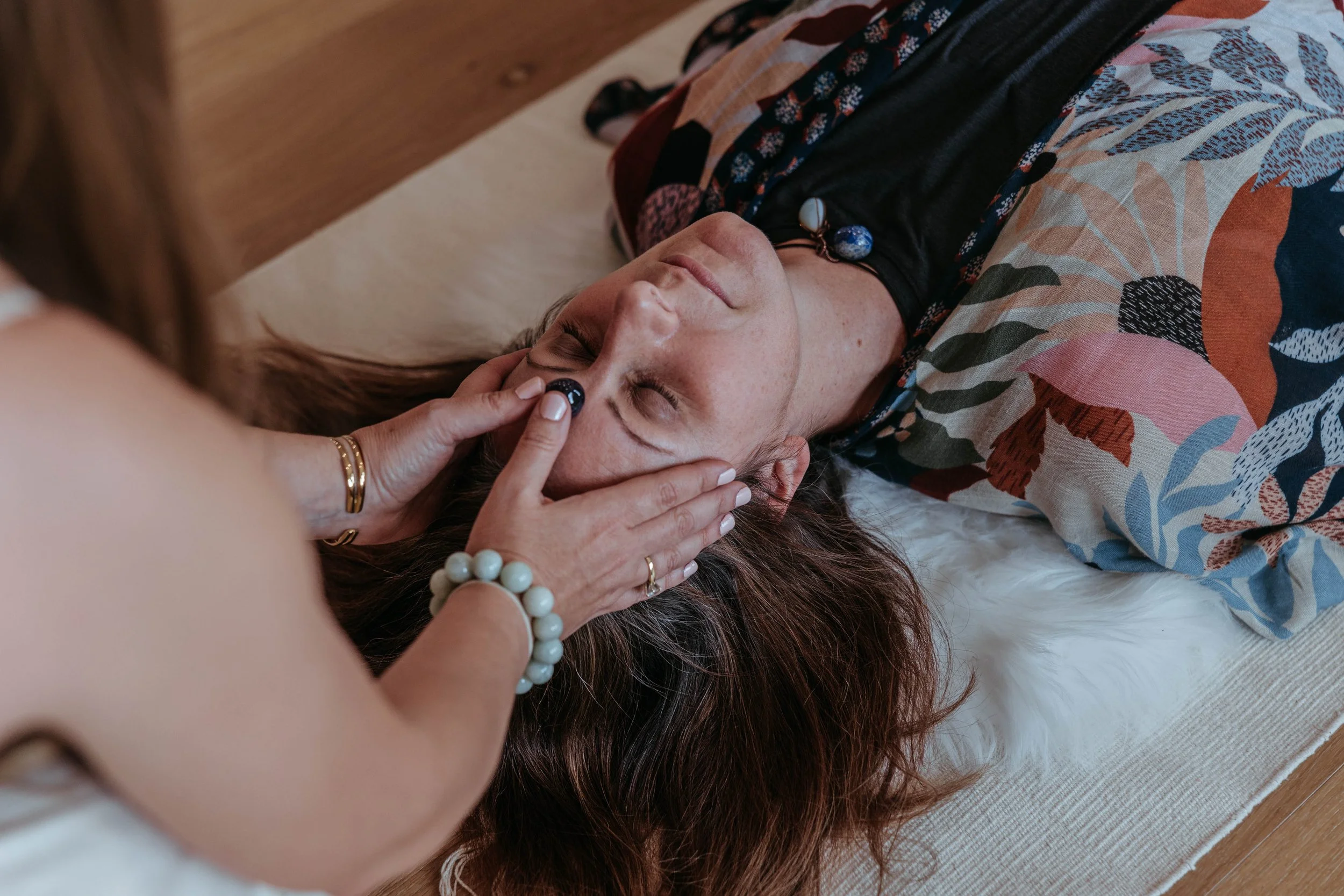 A woman lying on a mat with her eyes closed, receiving a facial massage or treatment from another woman using a black facial roller on her forehead.