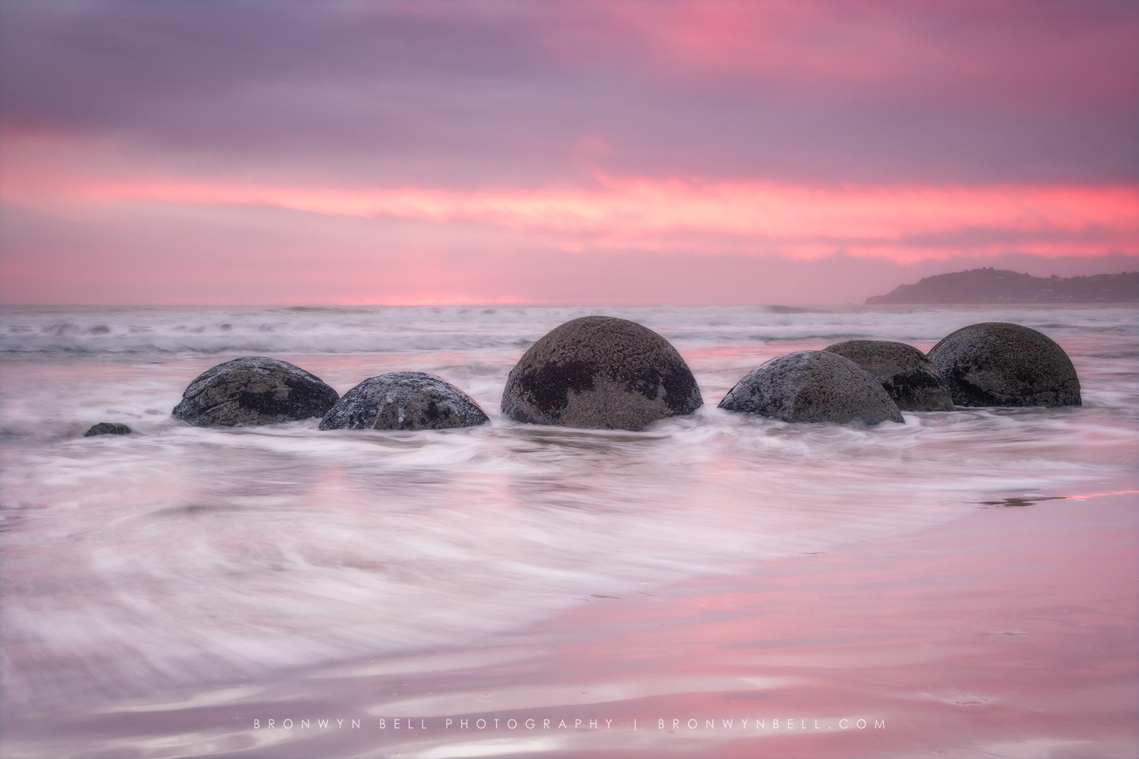 Beach with large round rocks partially submerged in ocean water during sunset with pink and purple sky, distant landmass on the horizon, and gentle waves.