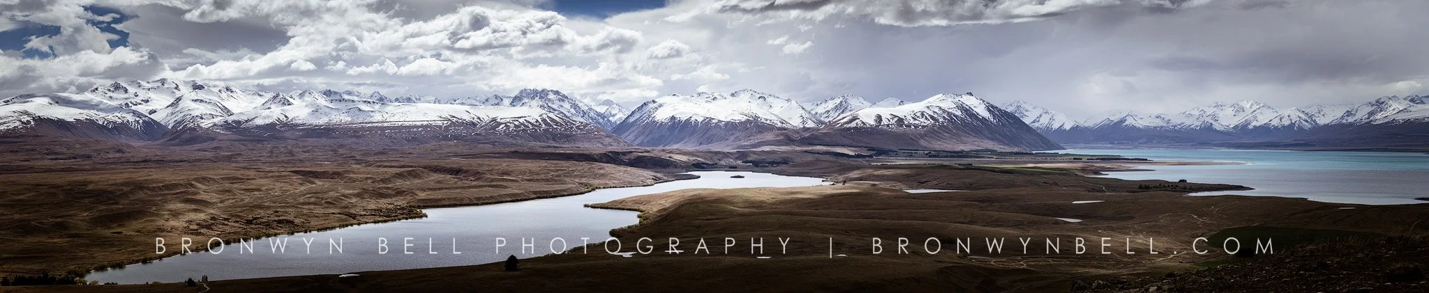 Scenic landscape of a winding river flowing through a brown grassy plain with snow-capped mountains in the background and partly cloudy sky.