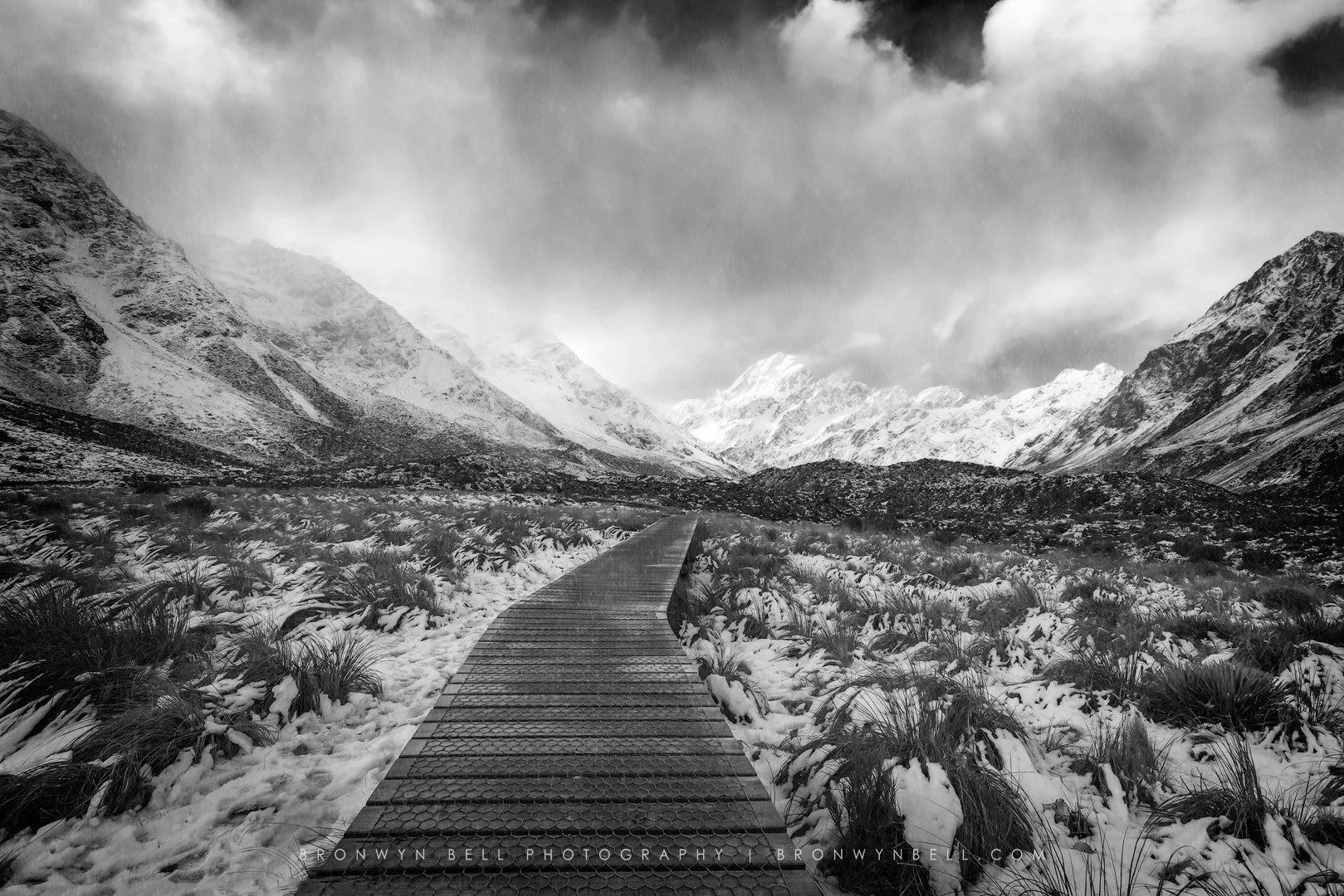 Snow-covered mountain valley with a wooden pathway leading towards distant snow-capped peaks under cloudy sky.