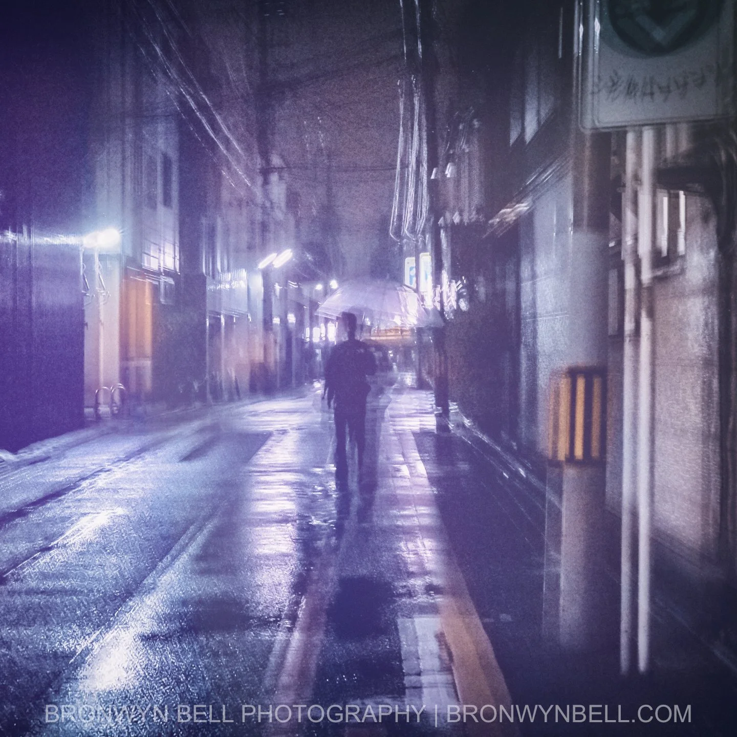 A lone figure draped in a dark coat walks under the dim glow of a corner light, the reflections on the wet pavement weaving into the soft edges of nearby buildings. The multi-exposure blurs time itself, letting the moment breathe with nostalgia.