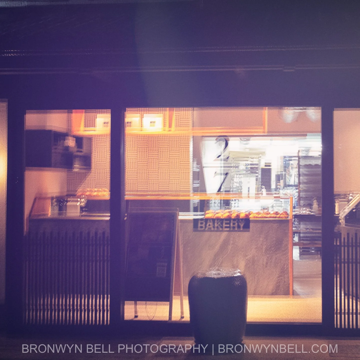 Through the misty rain of a Kyoto backstreet, the warm glow of a late-night bakery spills onto the slick pavement, reflecting in golden pools beneath tangled webs of overhead wires. Multi-exposures merge the soft outlines of pastry-filled shelves. An