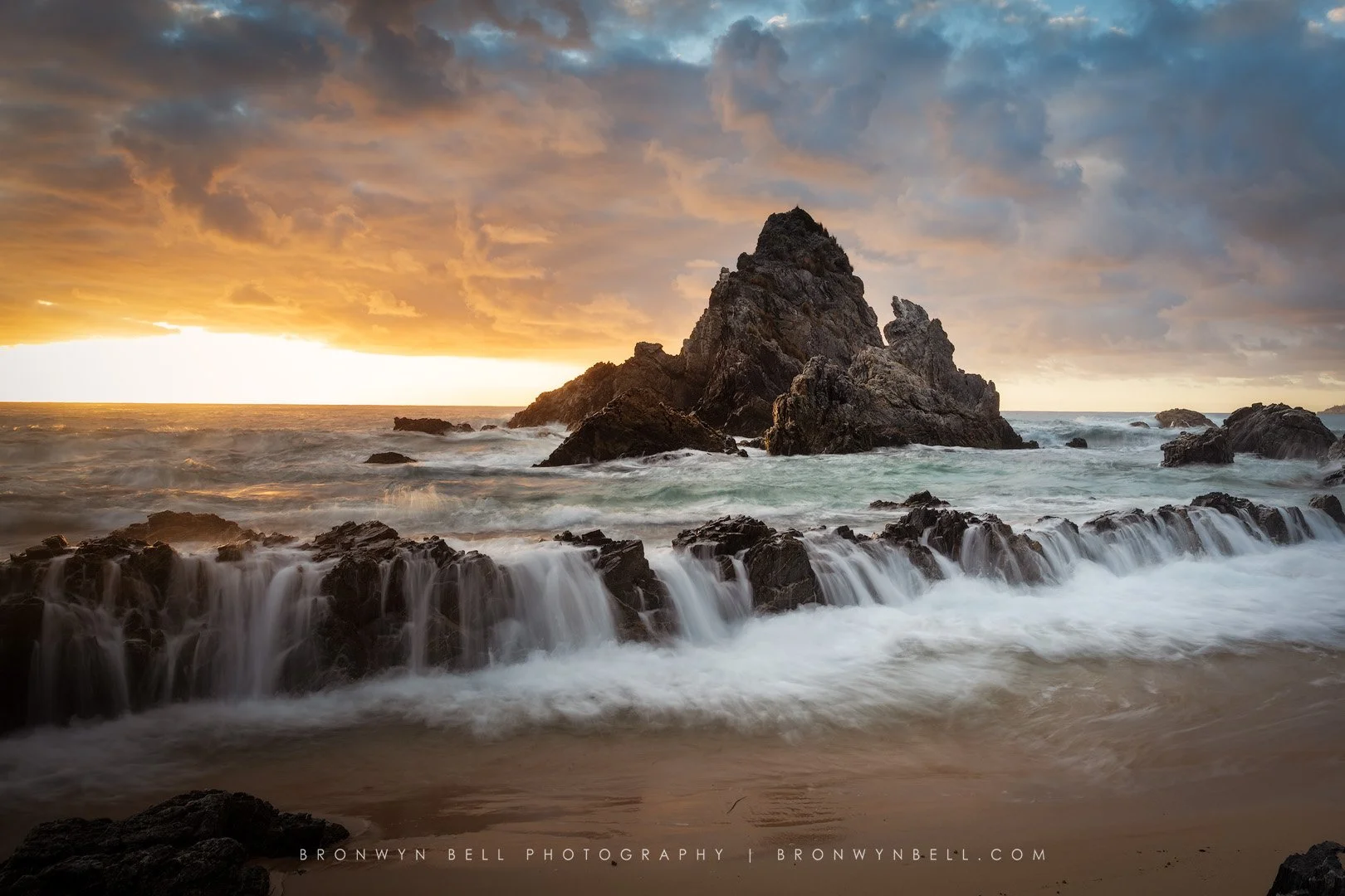 A rocky shoreline at sunset with waves crashing against the rocks and a large rock formation in the ocean, under a cloudy sky.