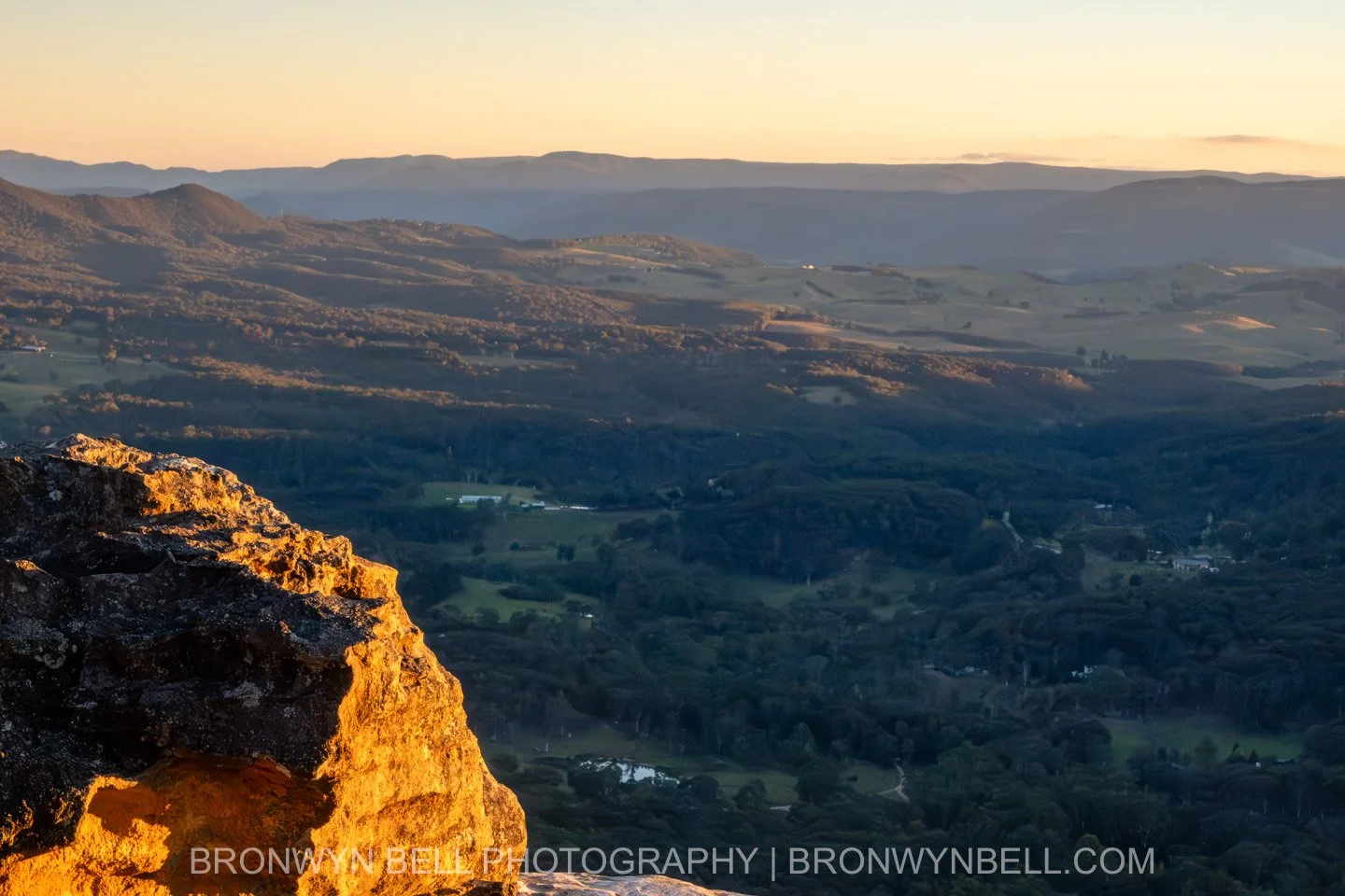 Landscape photograph from Sunset Rock in the Blue Mountains, with a large rocky outcrop in the foreground overlooking the Megalong Valley. The scene is lit by a warm sunset, with soft orange and golden tones in the sky.