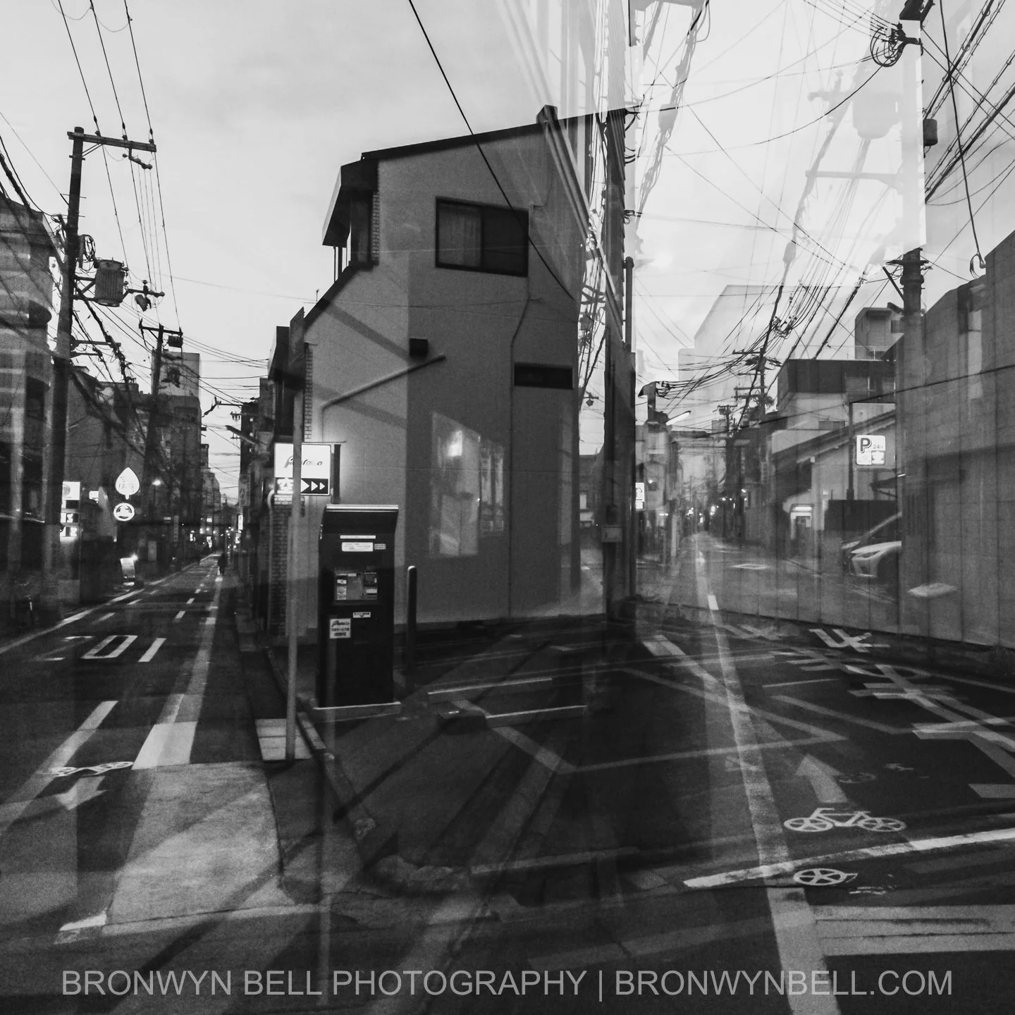 The crisscross of electricity wires cuts sharply against a pale sky, their angular lines mirrored by the roofs below. Layered exposures add depth to the weathered wood and stone, preserving the texture of centuries in black and white.