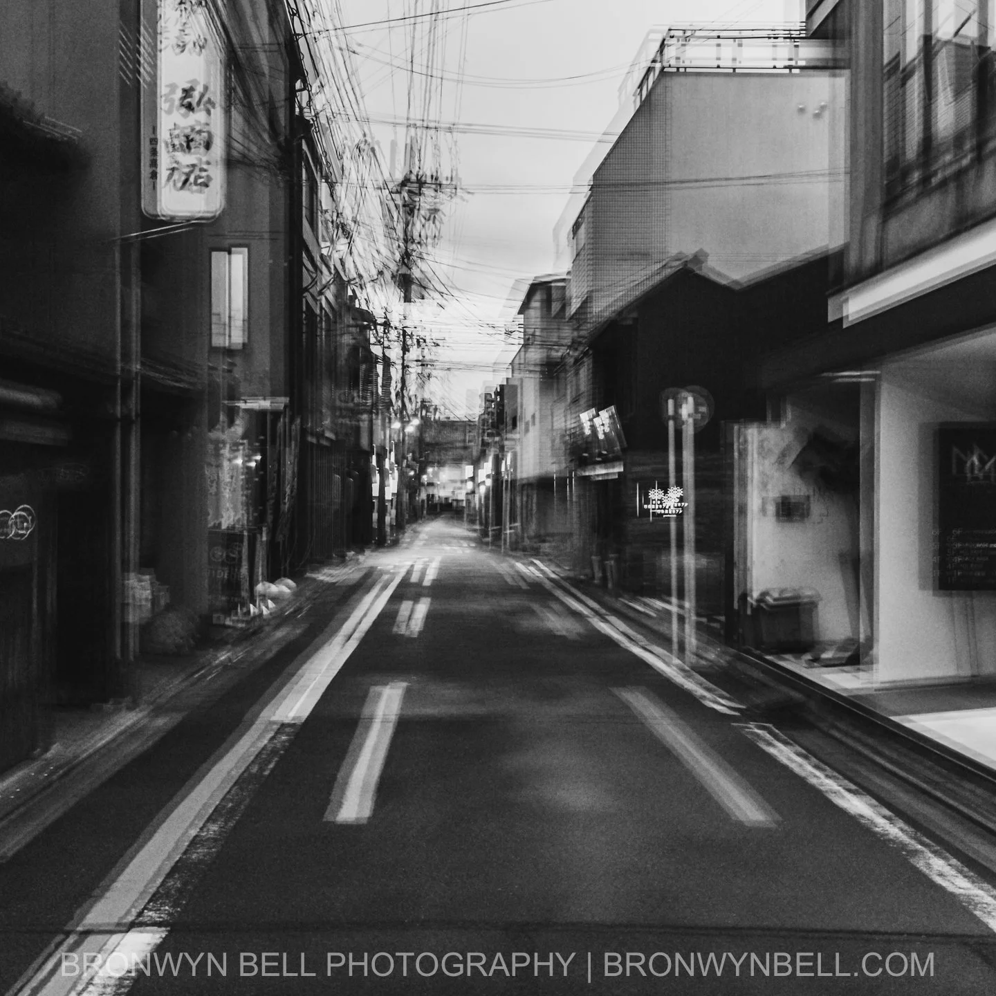 Morning light filters into a narrow lane, the close-set wooden façades leaning in like old friends. Multi-exposures blend rooftops, hanging cables, and shifting shadows into a timeless monochrome portrait of Kyoto’s quiet streets.