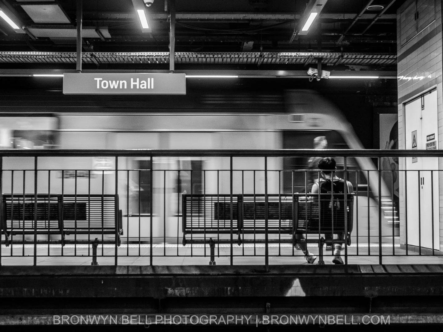 Black and white photograph of commuters waiting on the underground platform at Town Hall train station in Sydney while trains move through the station.