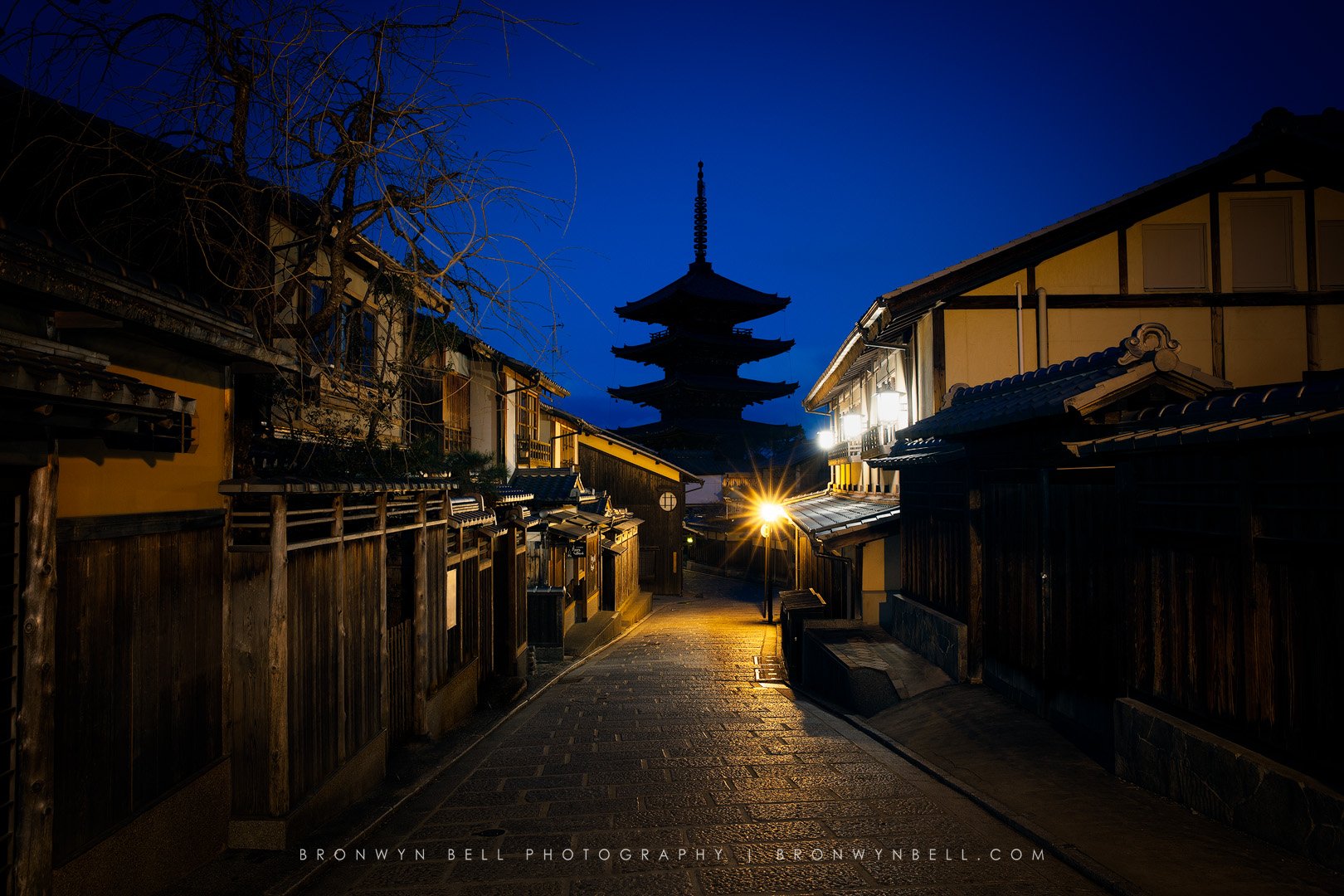Nighttime view of traditional Japanese street with wooden buildings and a pagoda in the background, illuminated by streetlights.