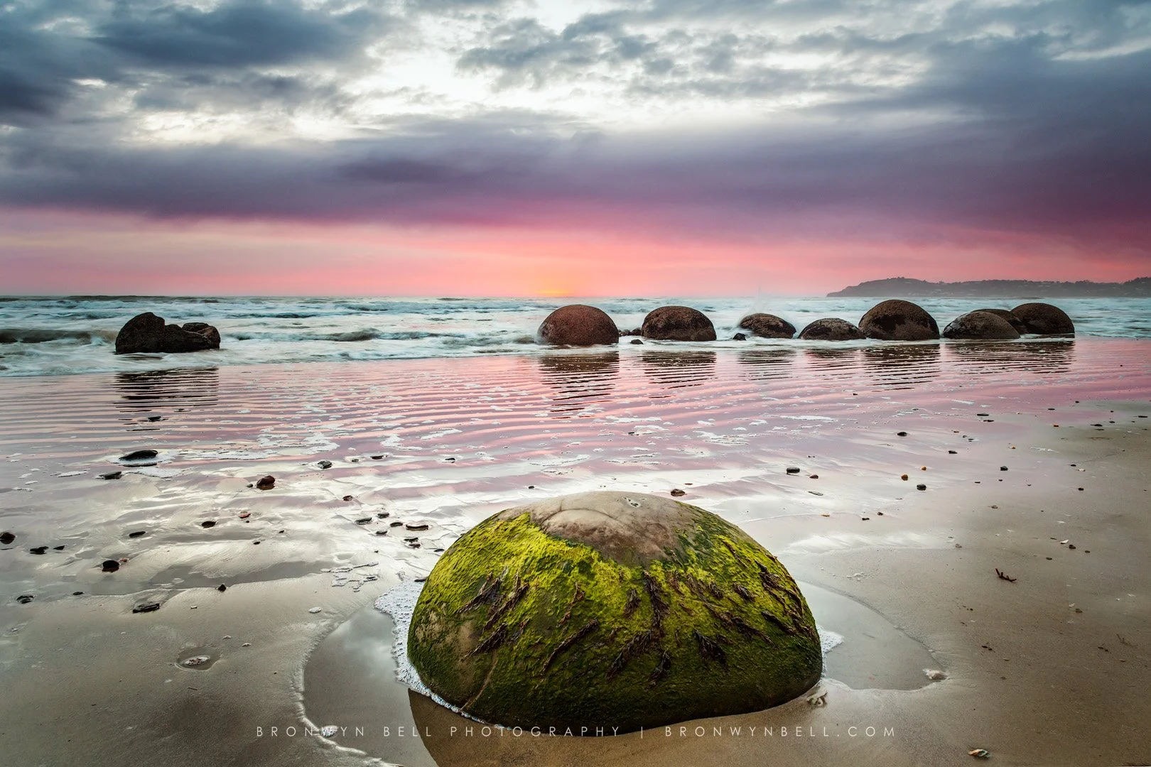 Moss-covered rock on beach with ocean and large rounded rocks in the background at sunset