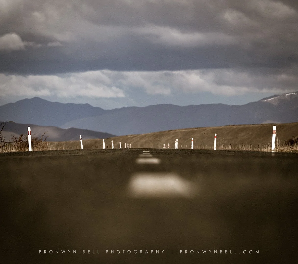 A long, empty road with white roadside posts stretches into the distance against a backdrop of mountains and a cloudy sky.
