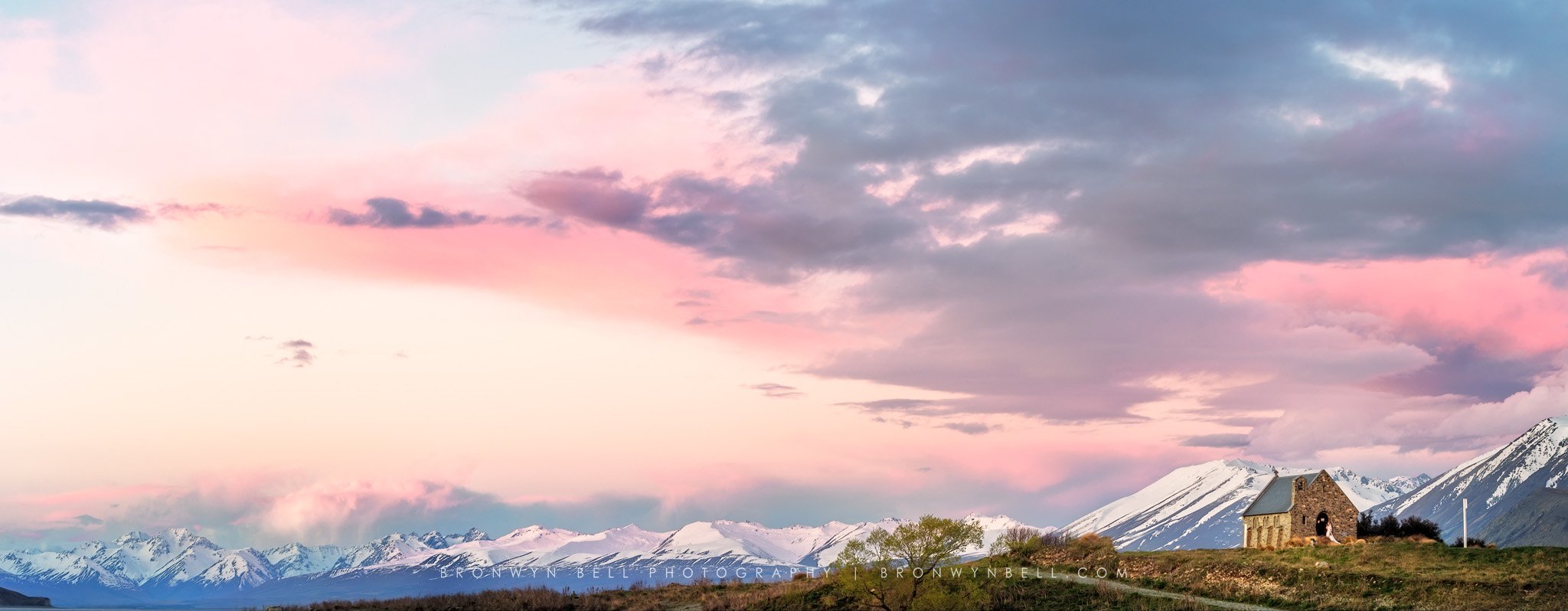 Scenic landscape with snow-capped mountains under a pastel pink and blue sky, a small stone church and a tree in the foreground.