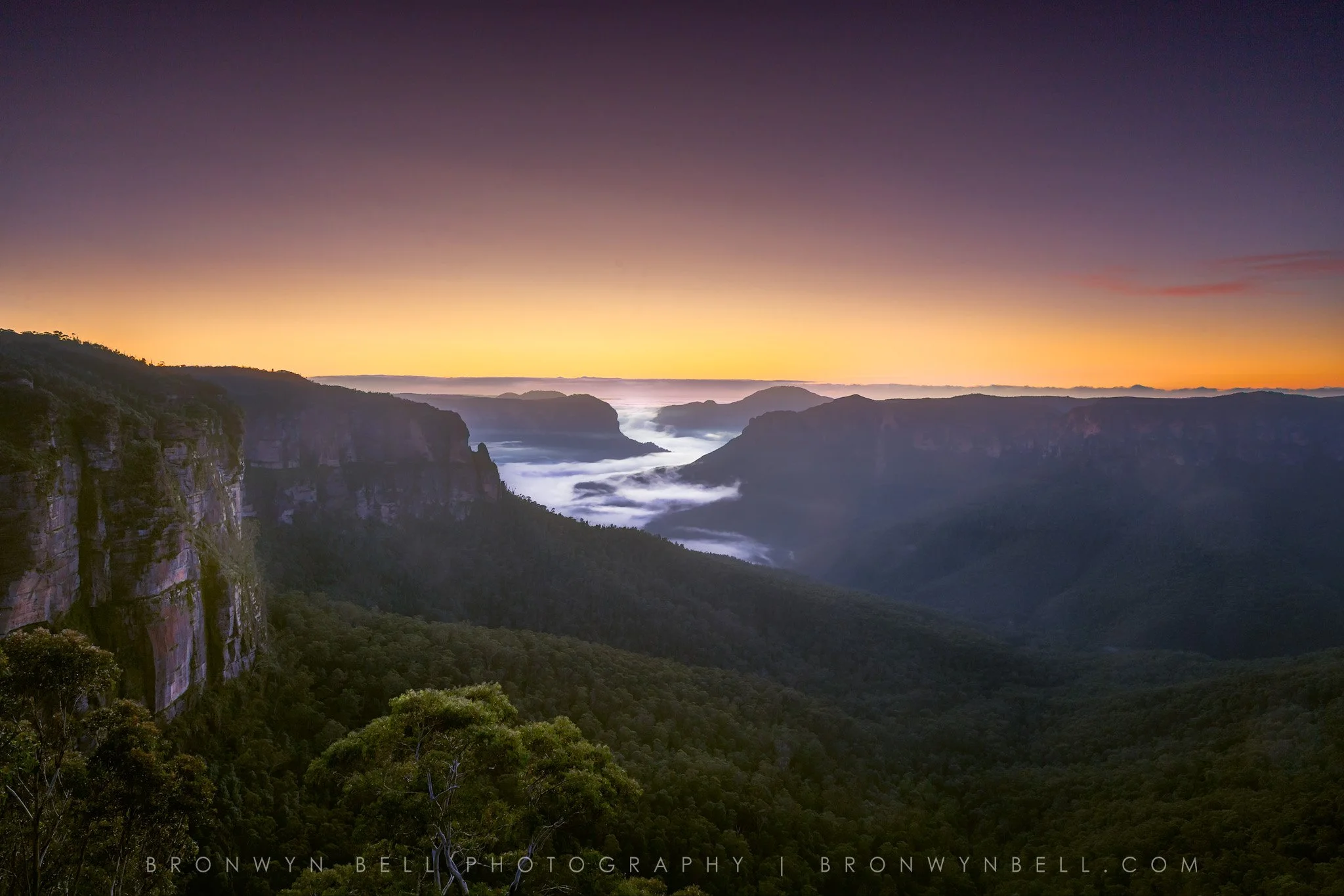 Sunrise over Govetts Leap is a breathtaking display of nature’s quiet drama—golden light spills across the horizon, casting deep orange and soft yellow hues into a sky awakening with warmth. Below, the Grose Valley is cloaked in mist, with rolling cl