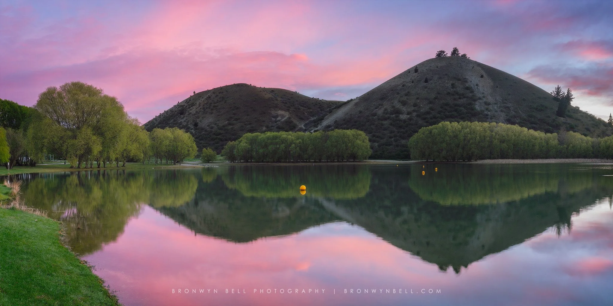 A serene lake reflecting surrounding green trees, hills with sparse vegetation, and a colorful pink and purple sky at sunset.