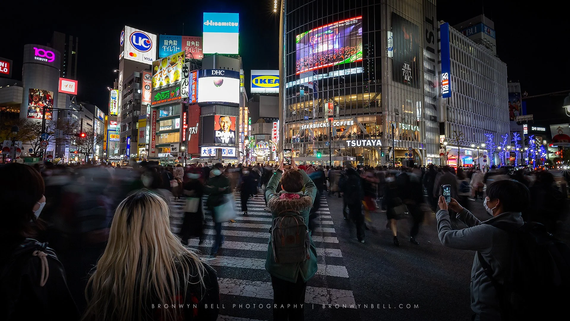Nighttime scene of a busy crosswalk in an urban area with brightly lit buildings and billboards, and people wearing masks crossing the street.