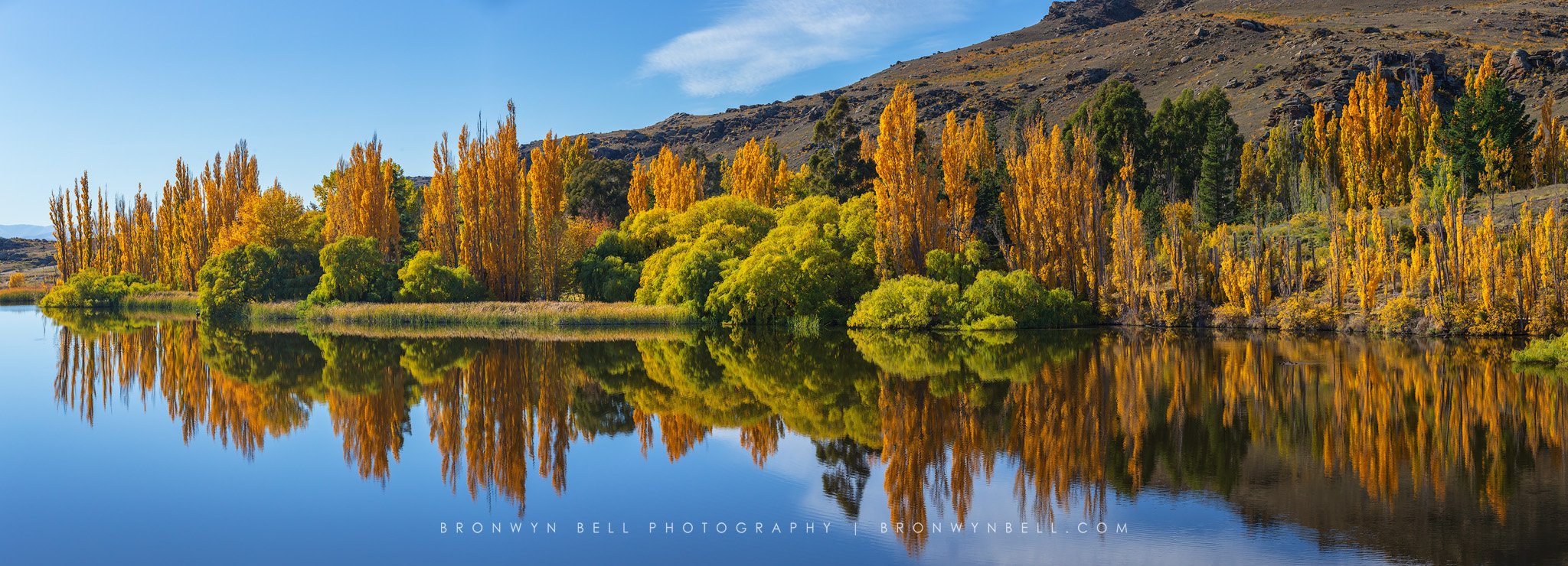 Autumn trees with yellow, orange, and green leaves reflected in a calm lake, mountains in the background, clear blue sky.