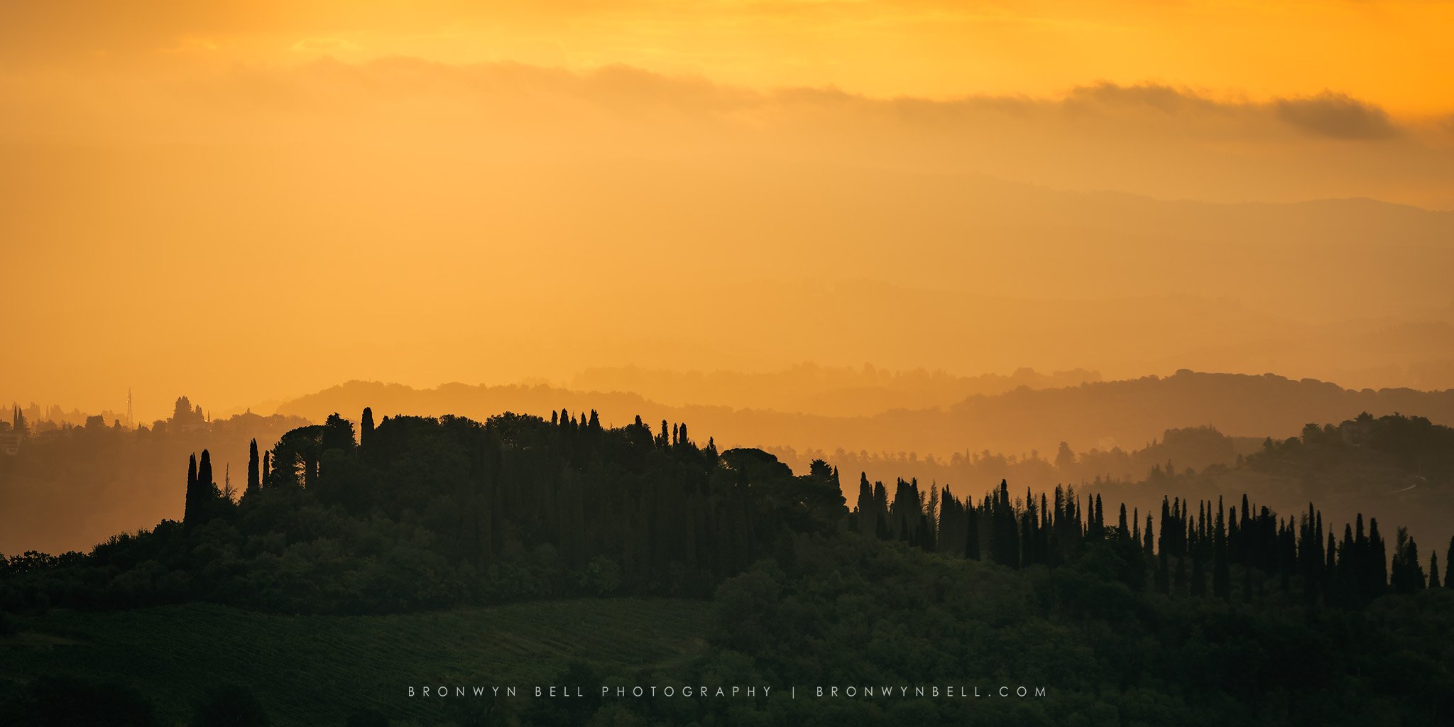Glowing orange sunset over the rolling hills of Tuscany, Italy, with dark green foreground, silhouetted pine trees and layered countryside fading into warm evening light in the distance