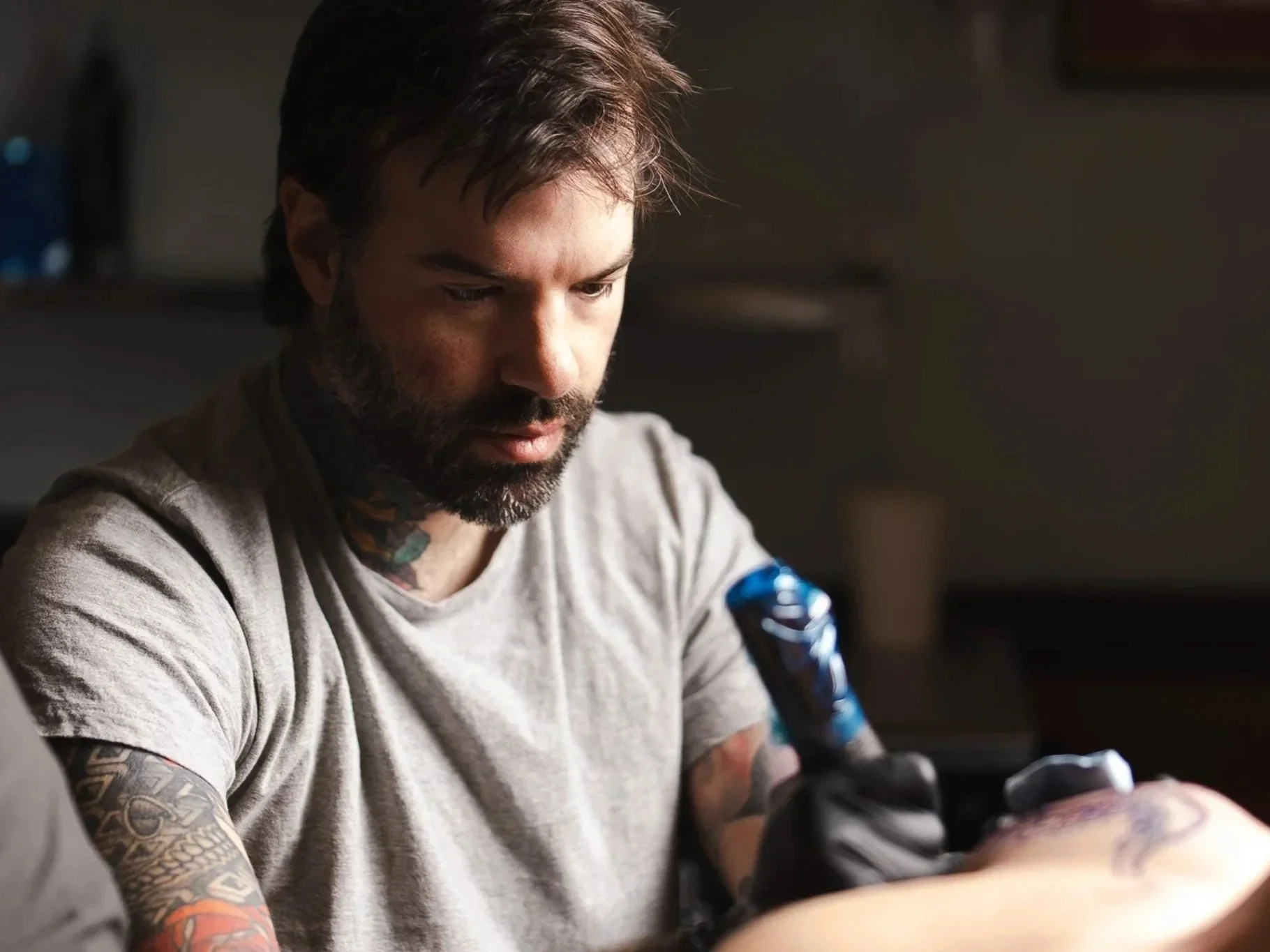 A man with dark hair and a beard tattooed on his neck and arms is tattooing another person's arm inside a dimly lit room.