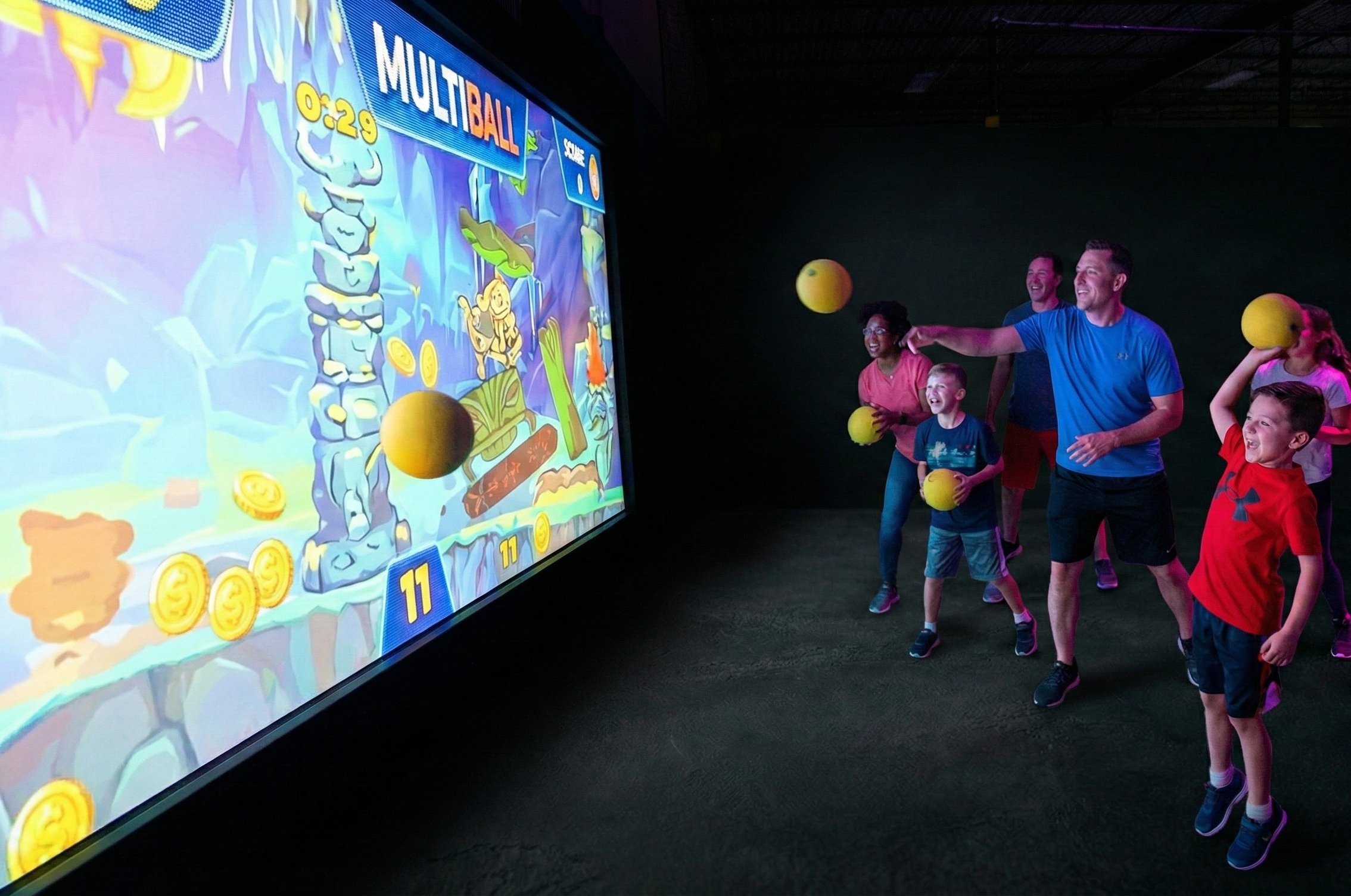 Group of children and adults playing a large arcade-style game involving hitting yellow balls at a screen showing a colorful game with time and scores.