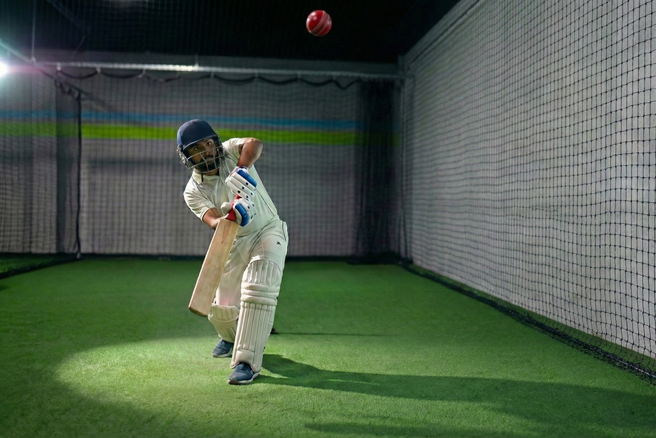 A cricket player batting in an indoor cricket net, wearing white uniform, helmet, and gloves, with a cricket ball headed toward him.