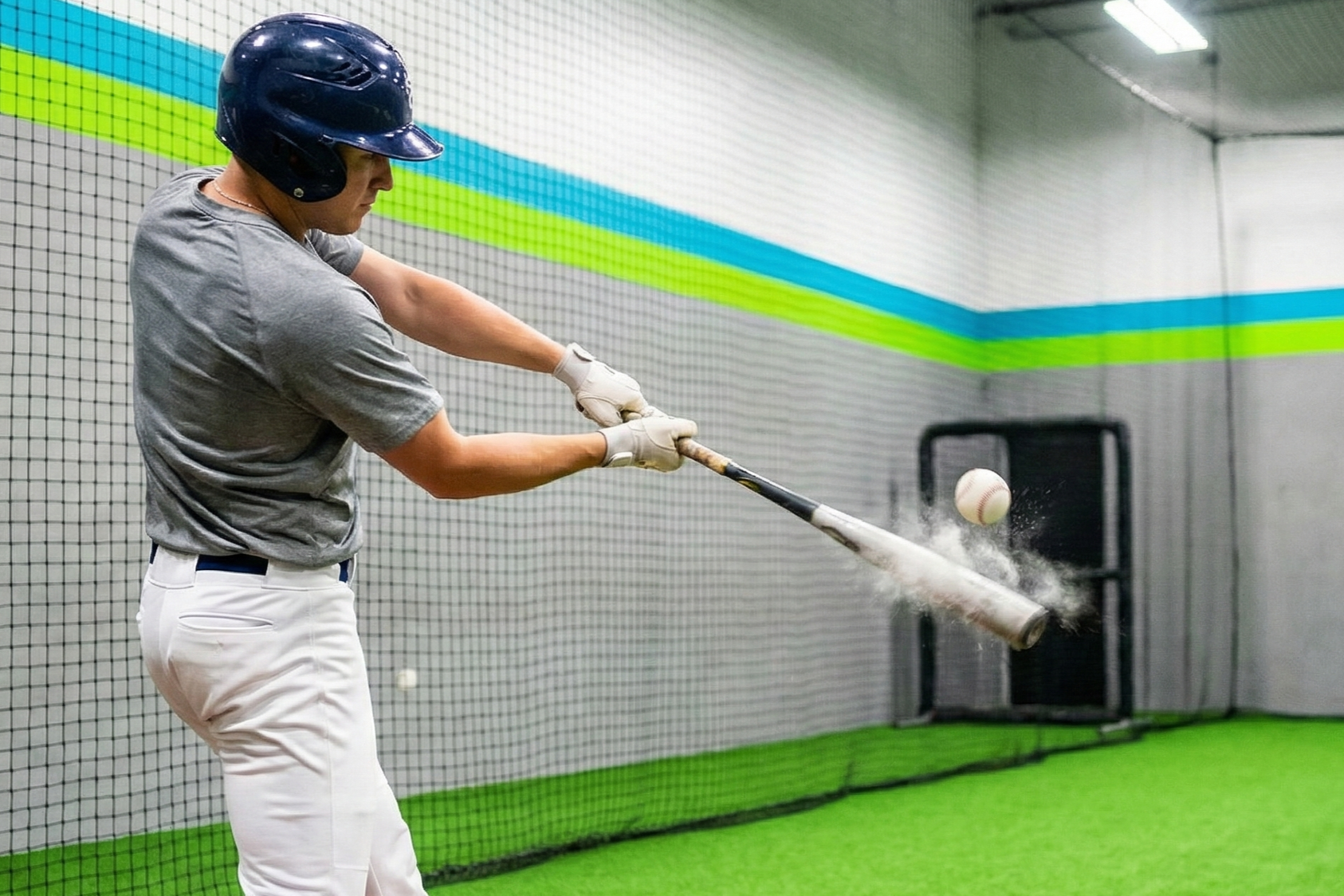 A young male baseball player practicing hitting in an indoor batting cage. He is wearing a navy helmet, gray shirt, white pants, and batting gloves. The baseball is in midair as he swings a bat, making contact with the ball.