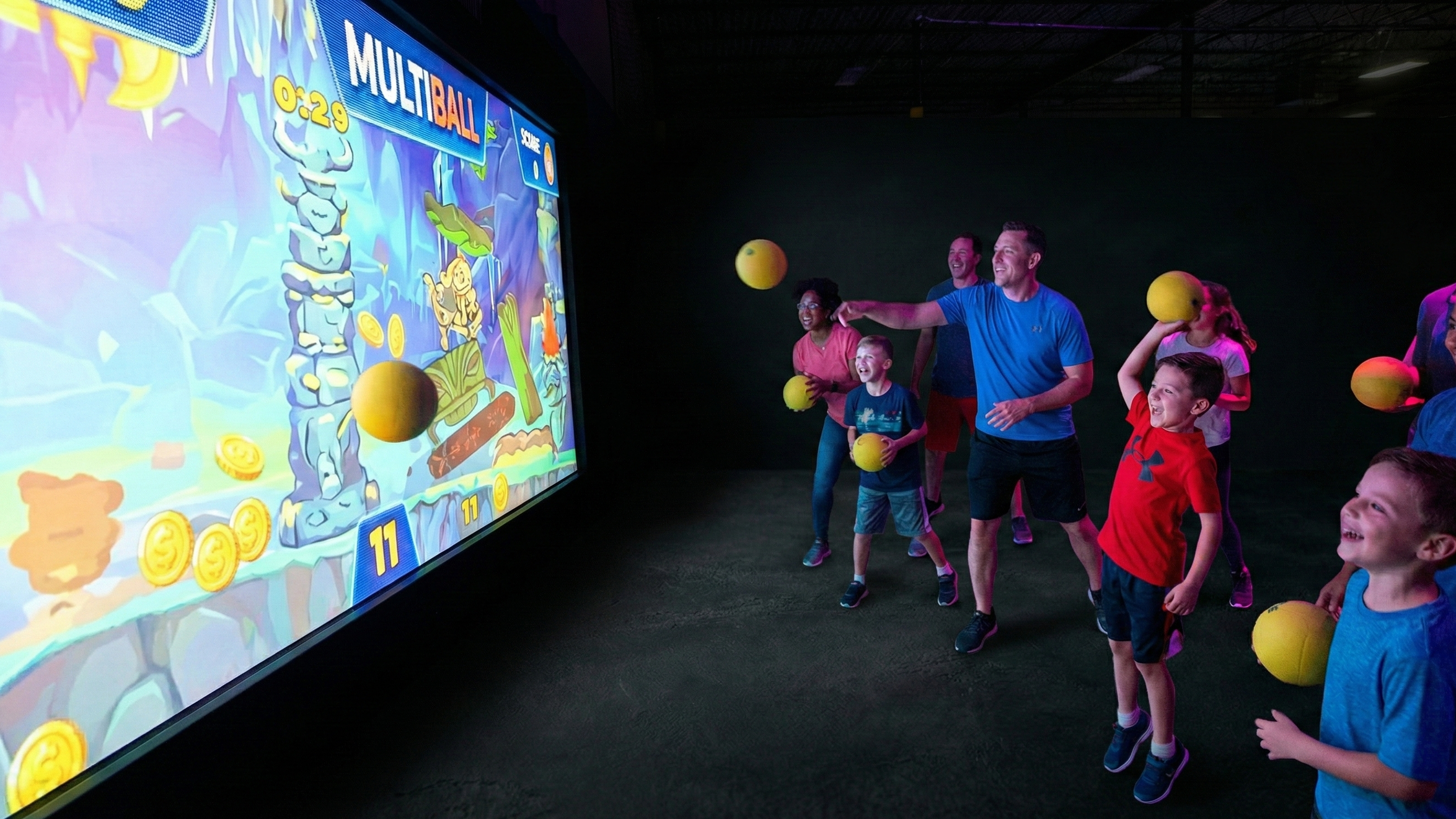 People, both children and adults, playing an interactive arcade game on a large screen, with some holding yellow balls and smiling as they take turns hitting the screen during a fun activity.