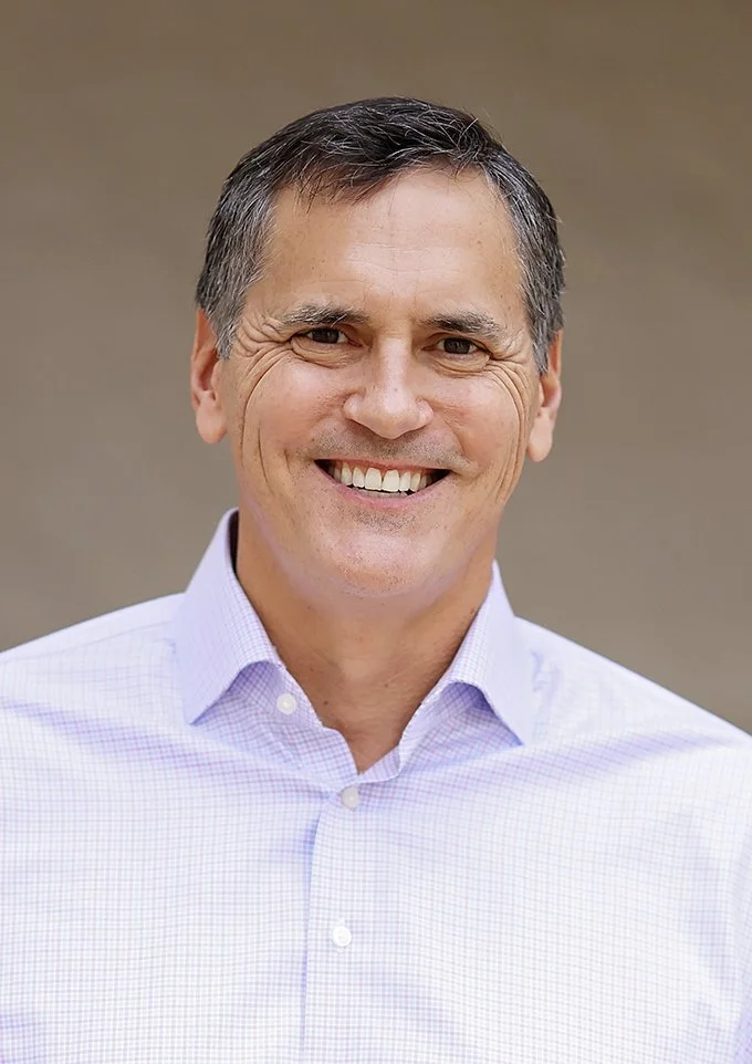 Portrait of a middle-aged man with short dark hair, smiling, wearing a light purple checkered dress shirt, standing outdoors against a neutral background.
