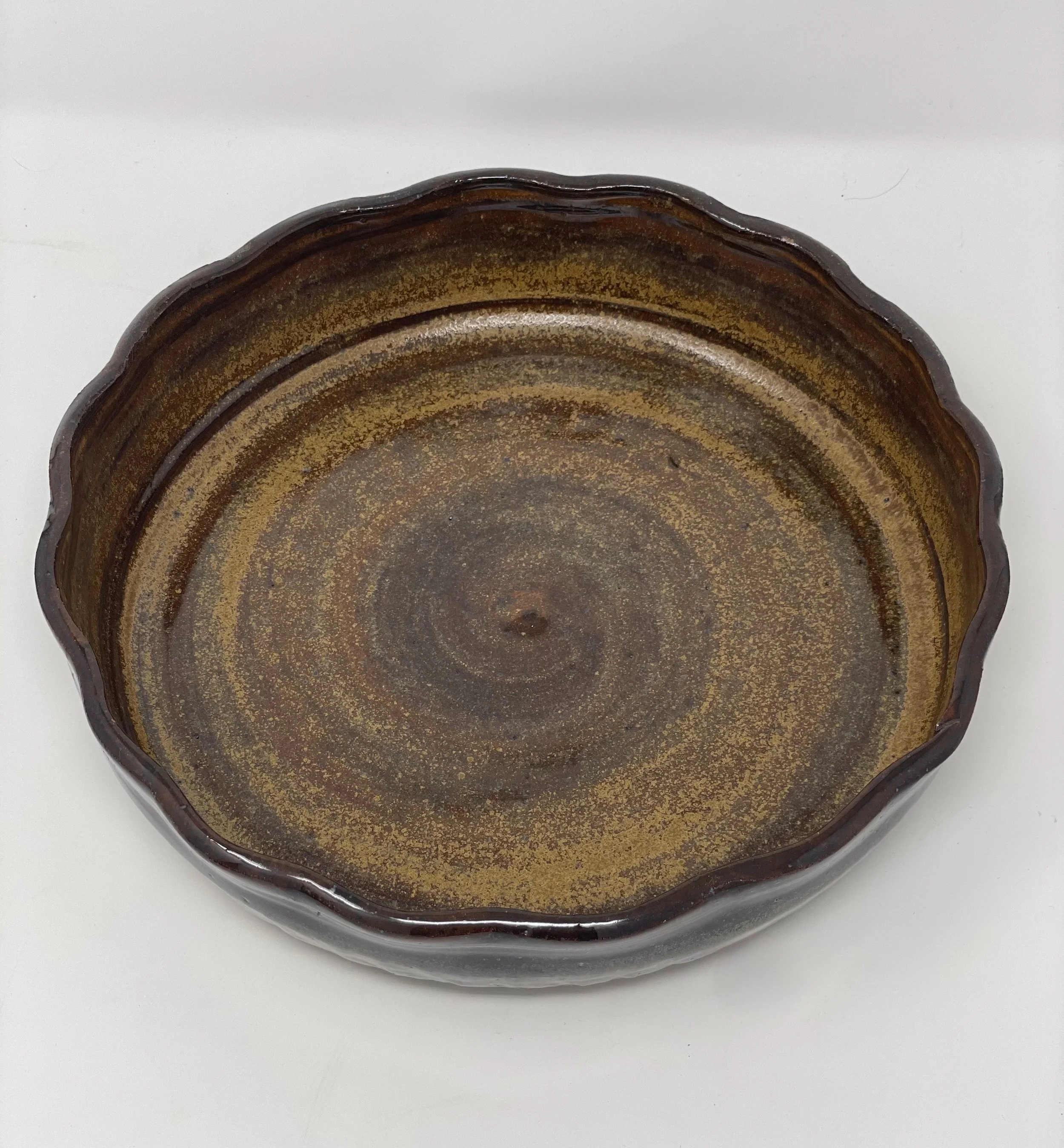 A circular, wavy-edged, vintage ceramic bowl with a brown and gold glazed interior, placed on a white surface.