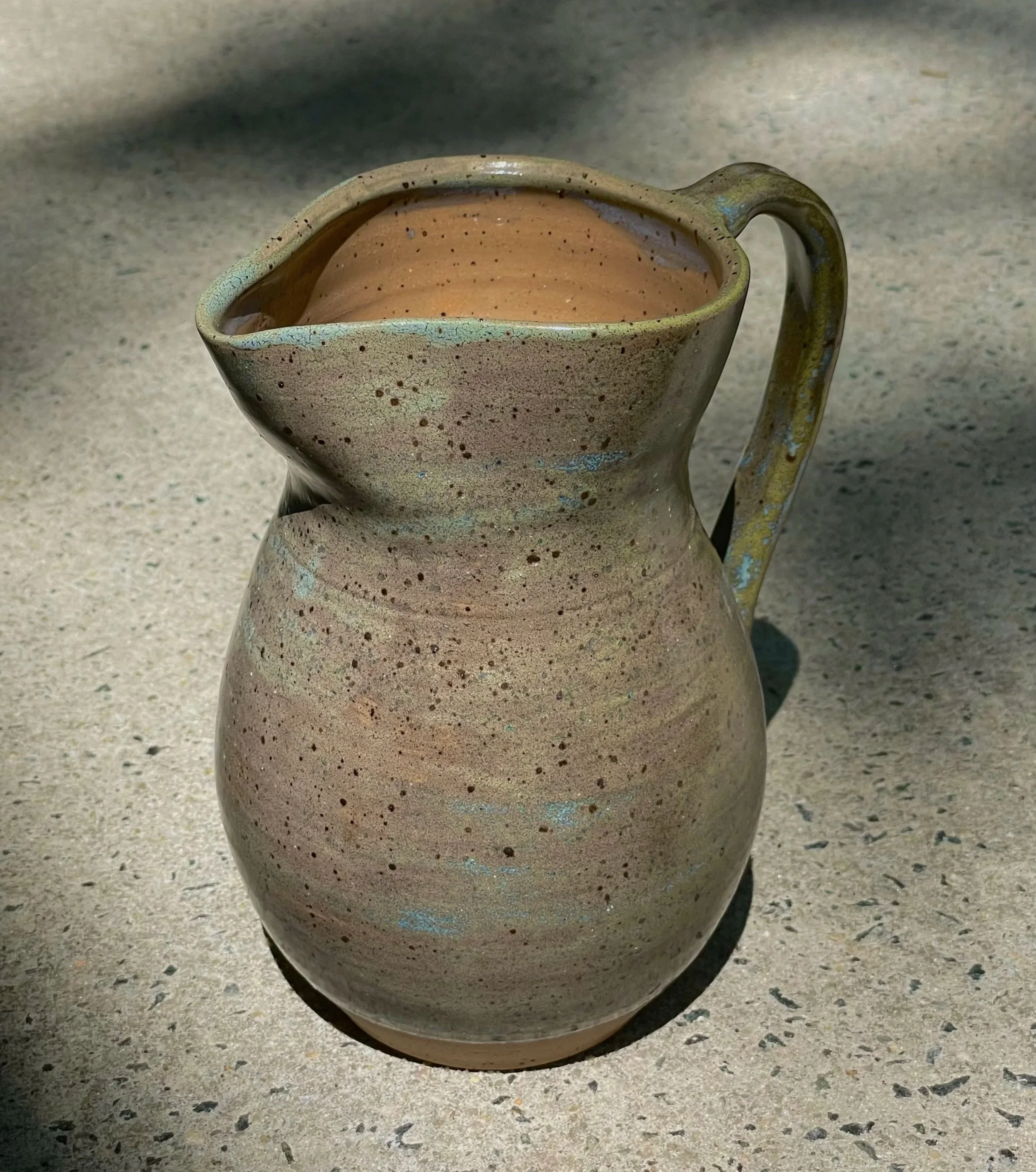 A ceramic pitcher with a speckled glaze, placed on a concrete surface with shadows.