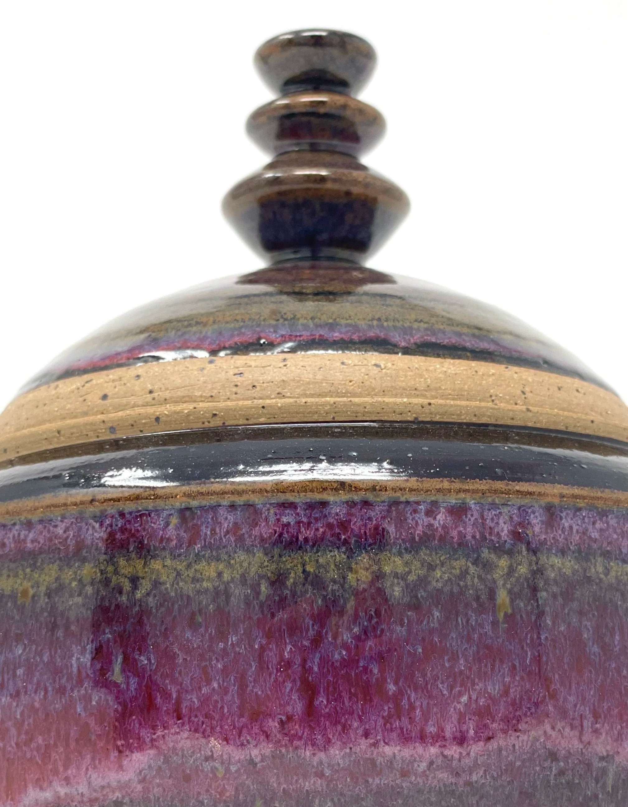 Close-up of a decorative ceramic piece with horizontal ridges and a spire on top, featuring black, purple, and earthy tones.