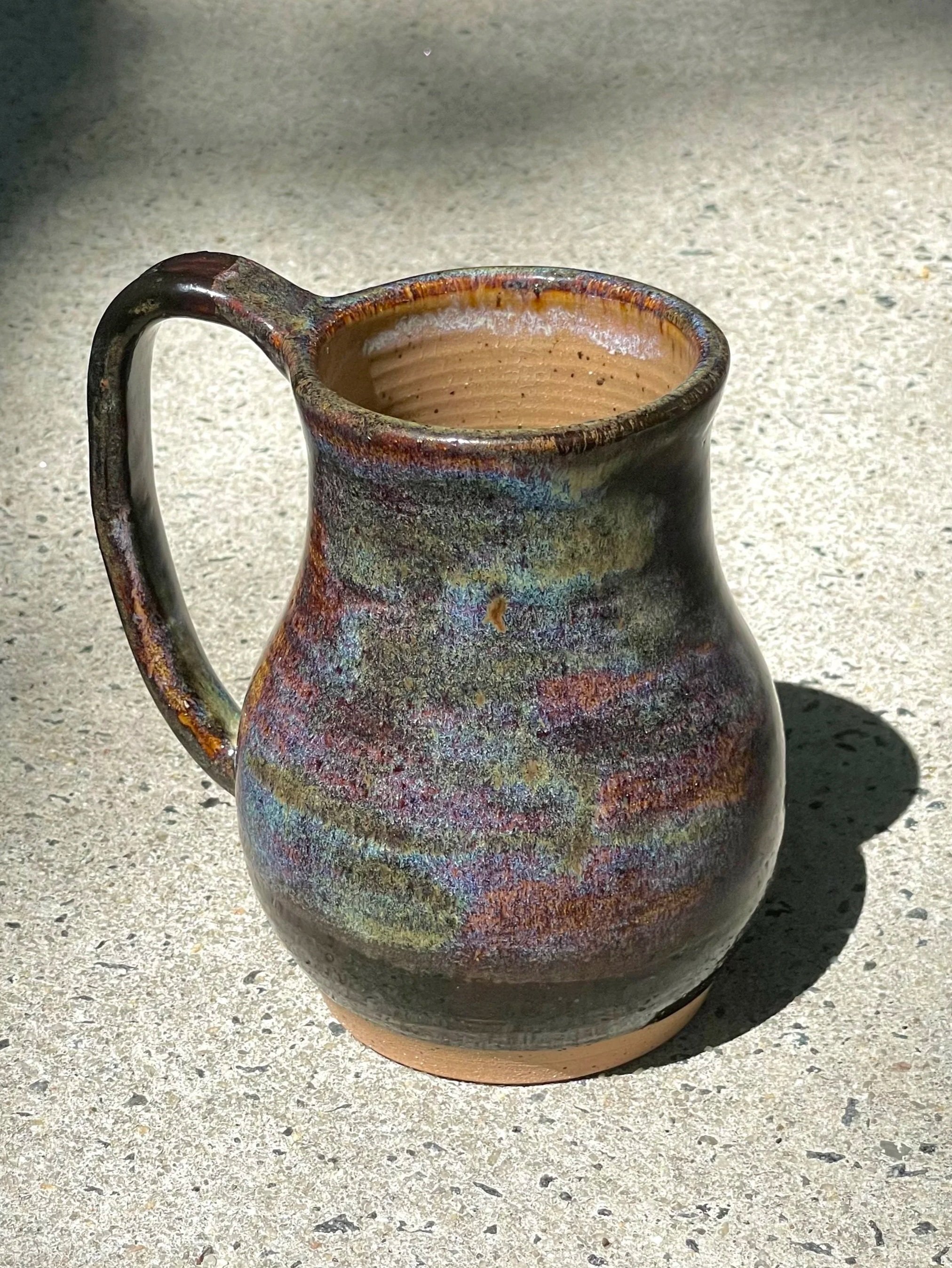 A ceramic mug with a multicolored, glossy glaze on the outside, sitting on a concrete surface in sunlight, with a shadow cast to the right.