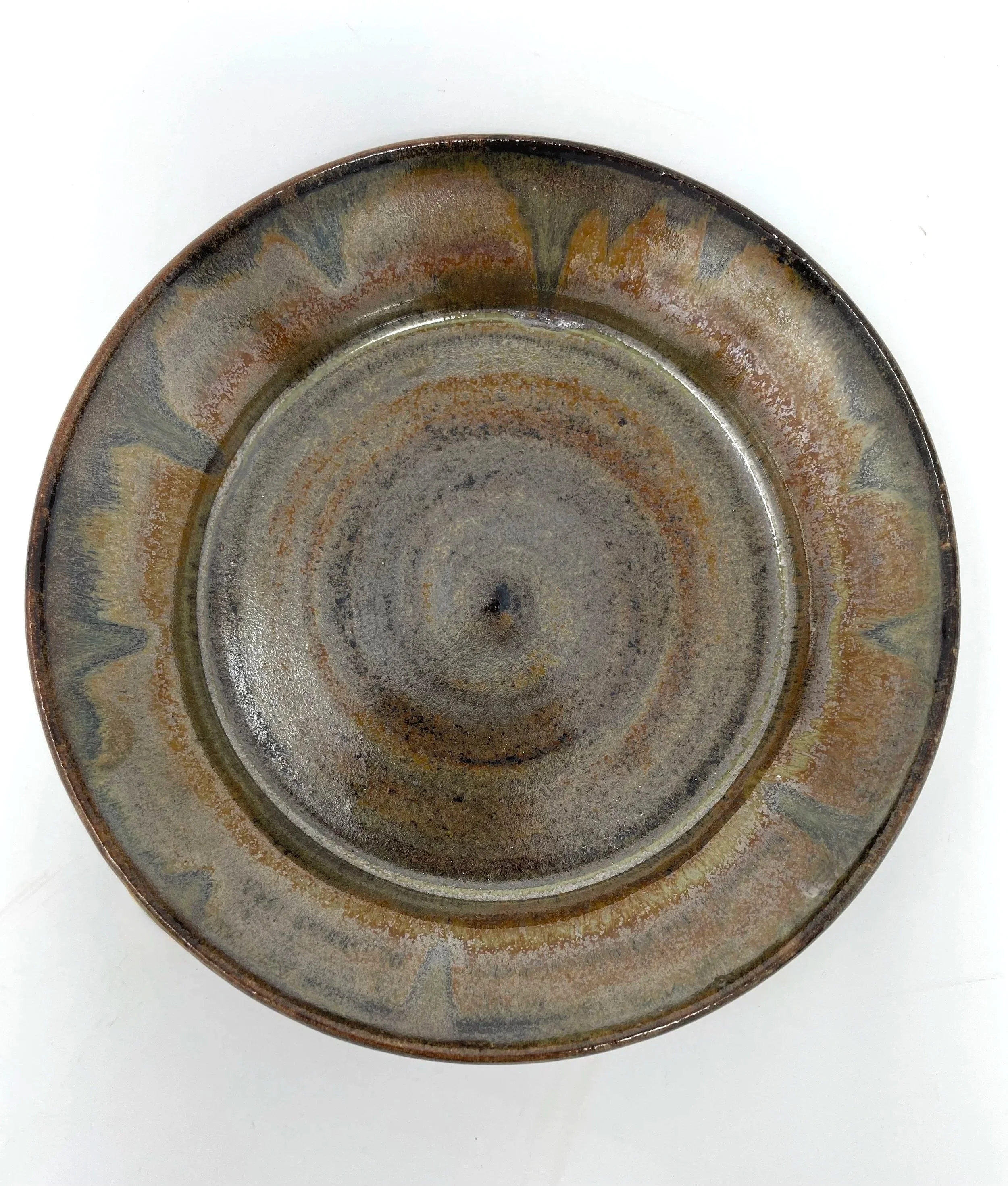 A round ceramic plate with a glazed, textured surface and earthy tones, featuring a decorative pattern around the rim with colors like brown, gray, and hints of yellow, set against a white background.