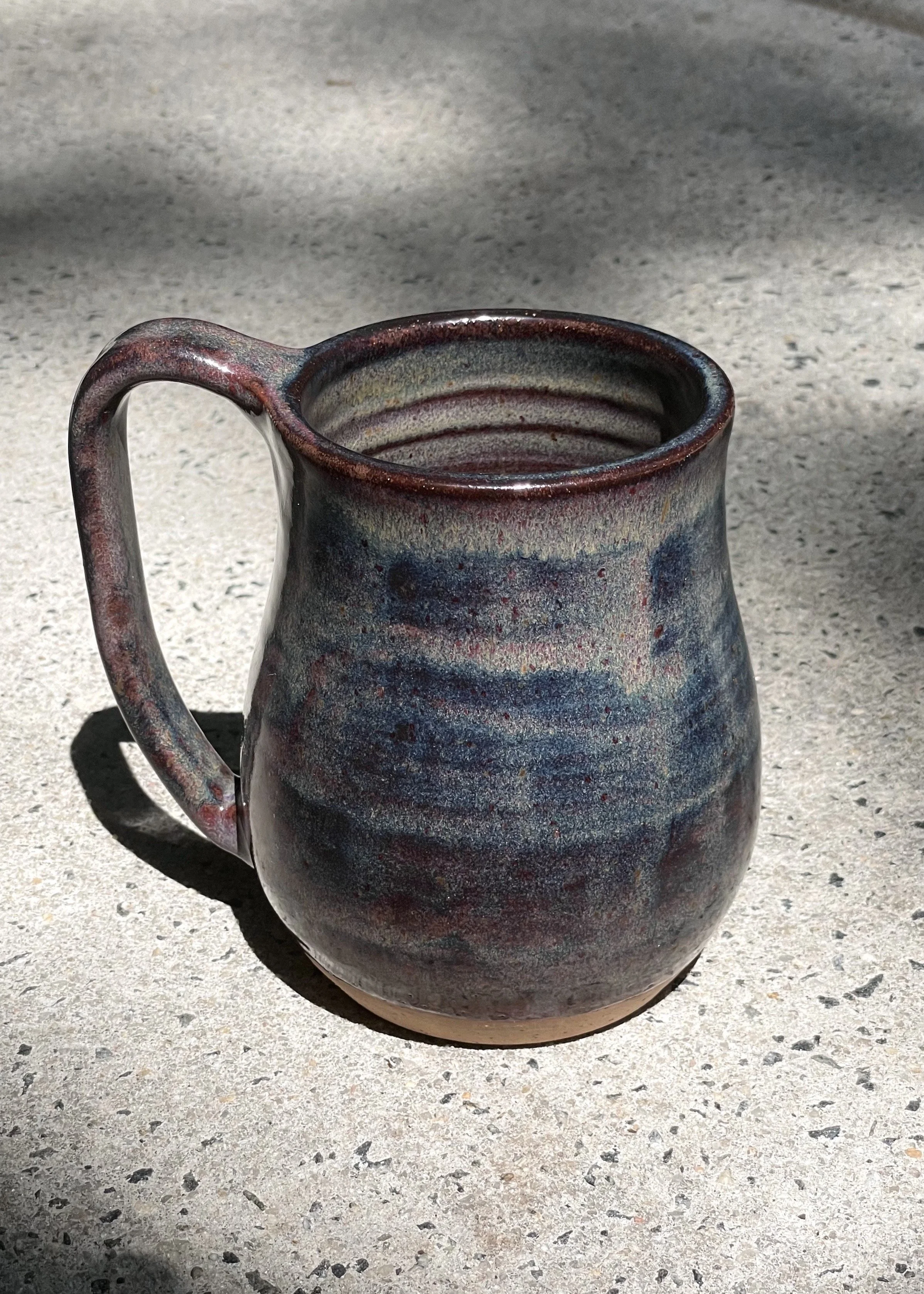 Ceramic mug with a multicolored glaze, placed on a concrete surface with sunlight and shadow.