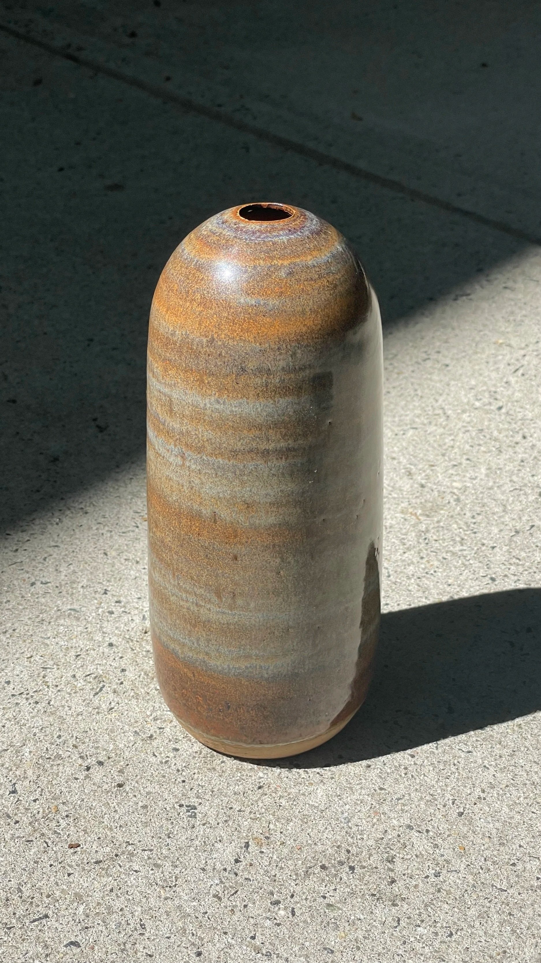 A tall sandy colored ceramic vase with a rounded top and narrow opening, featuring earthy-toned horizontal stripes, placed on concrete ground with shadows.