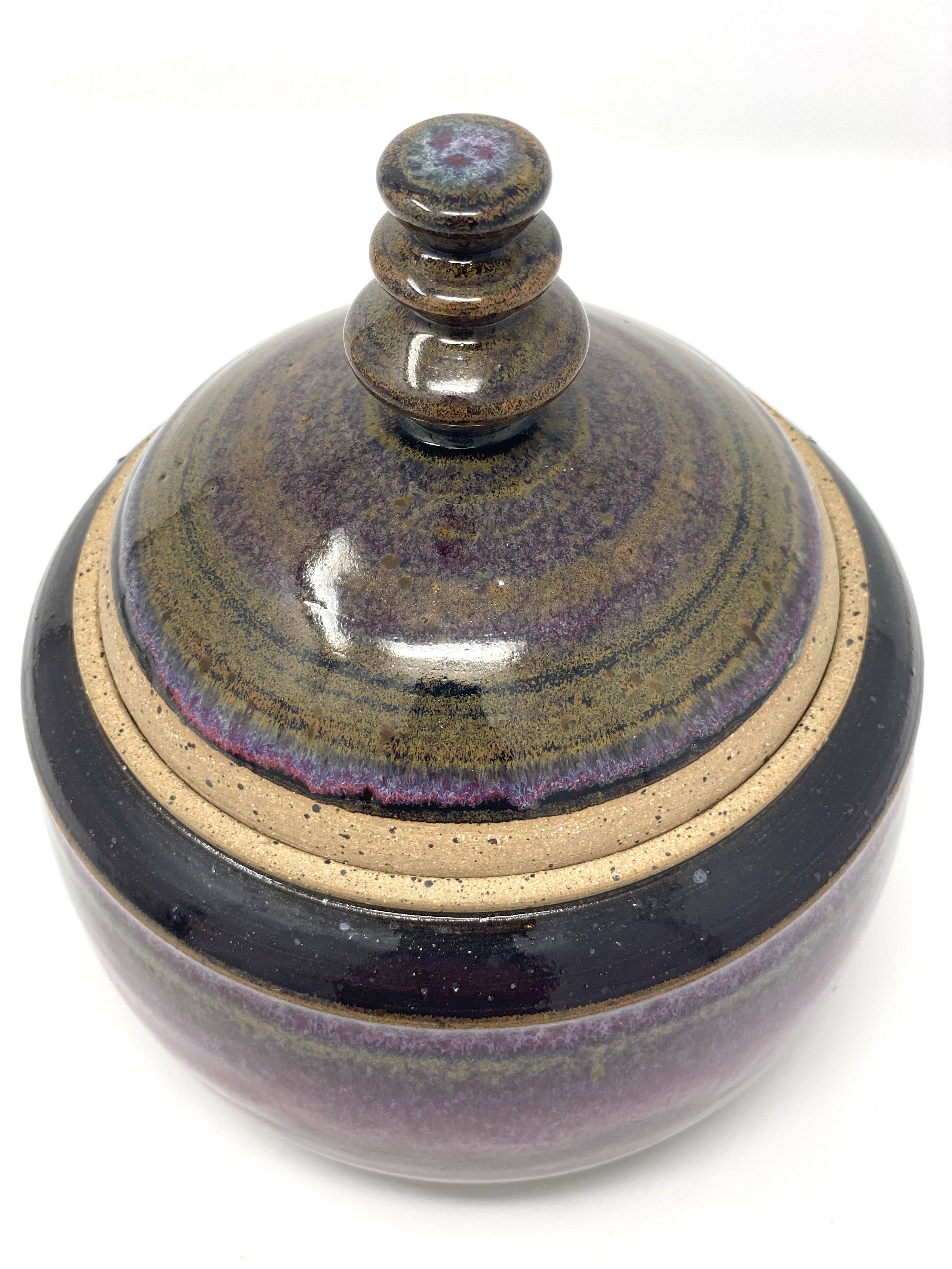 Close-up of a ceramic lid with a stacked knob handle, glazed in metallic and earthy tones with purple and gold accents.