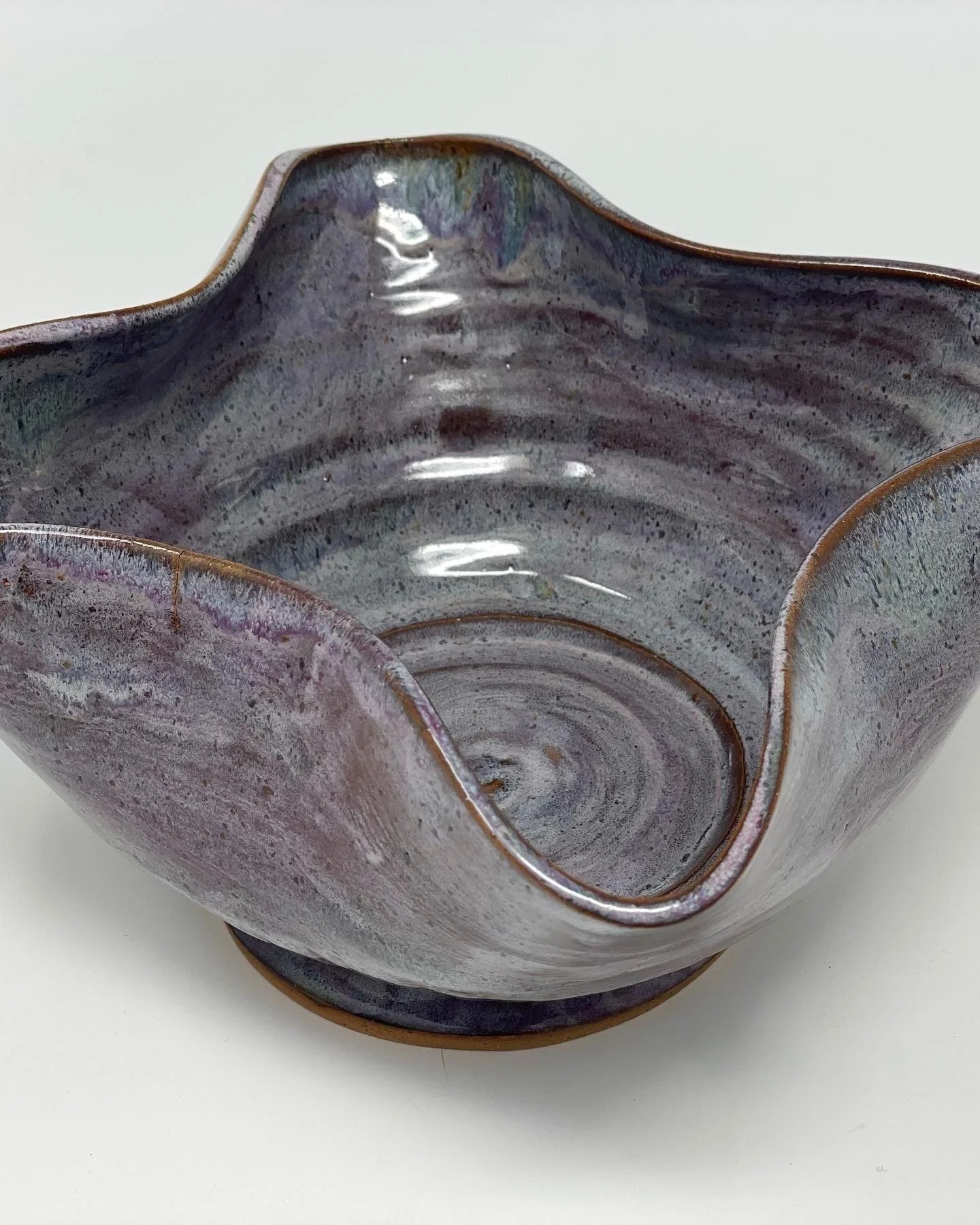 A handmade ceramic bowl with a wavy, petal-like edge, glazed in a mix of muted purple, blue, and gray tones, with a glossy finish and subtle swirl patterns inside.