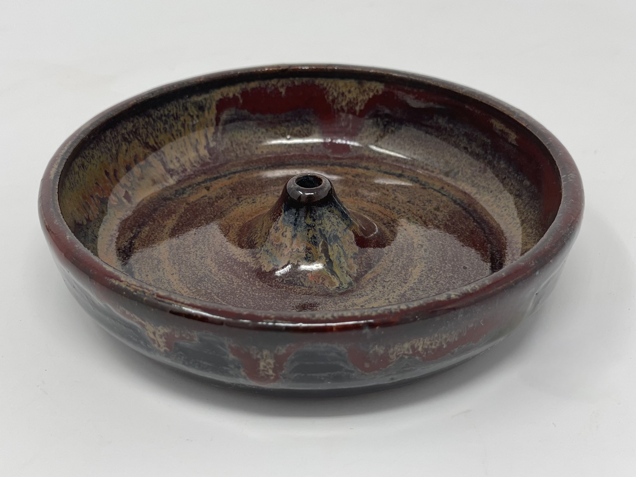 A round ceramic incense holder with a colorful glaze pattern containing dark, earthy tones and a small opening in the center for holding incense.