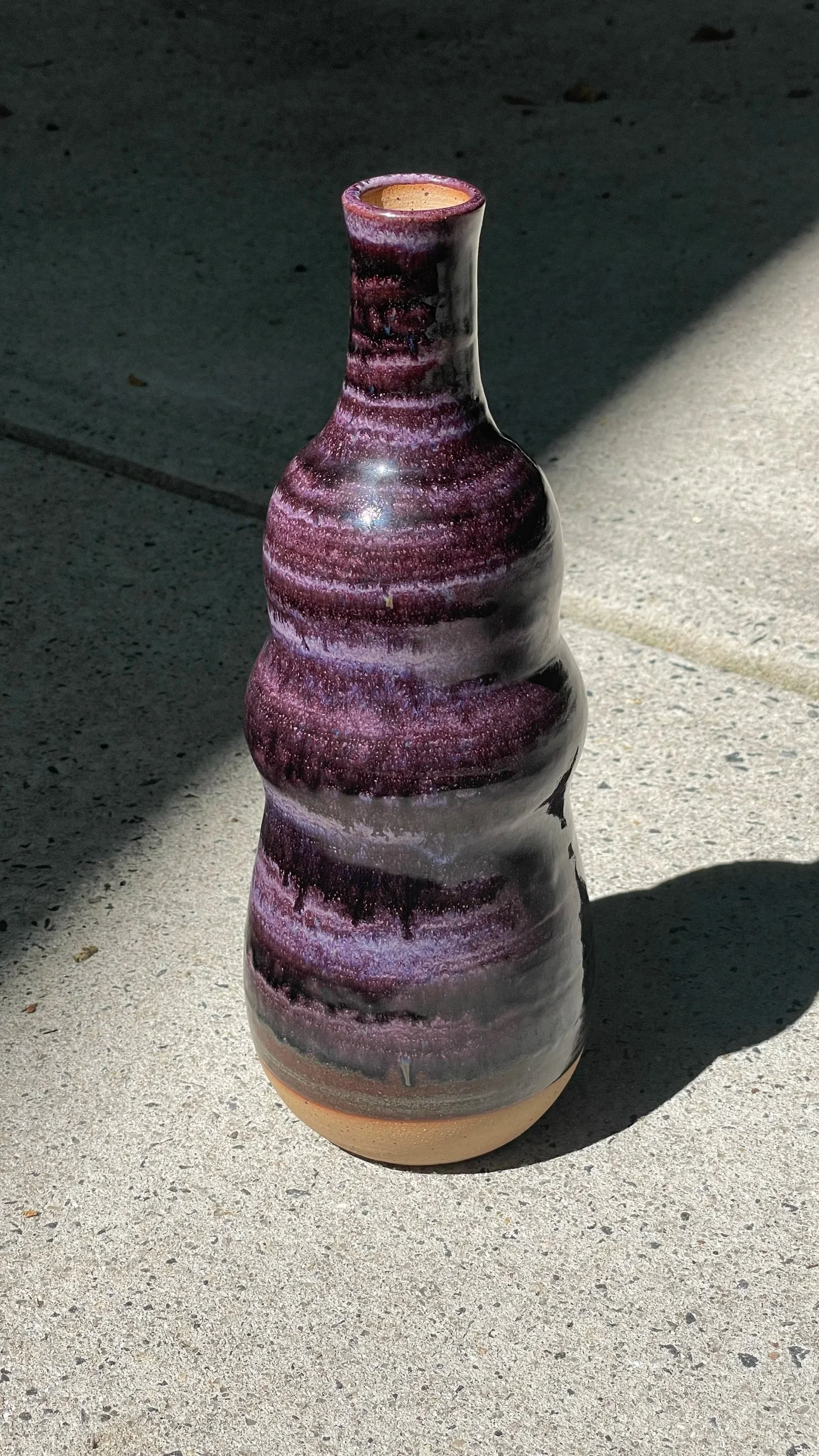 A tall, purple ceramic vase with shades of purple, black, and beige, standing on a concrete surface with sunlight casting a shadow.
