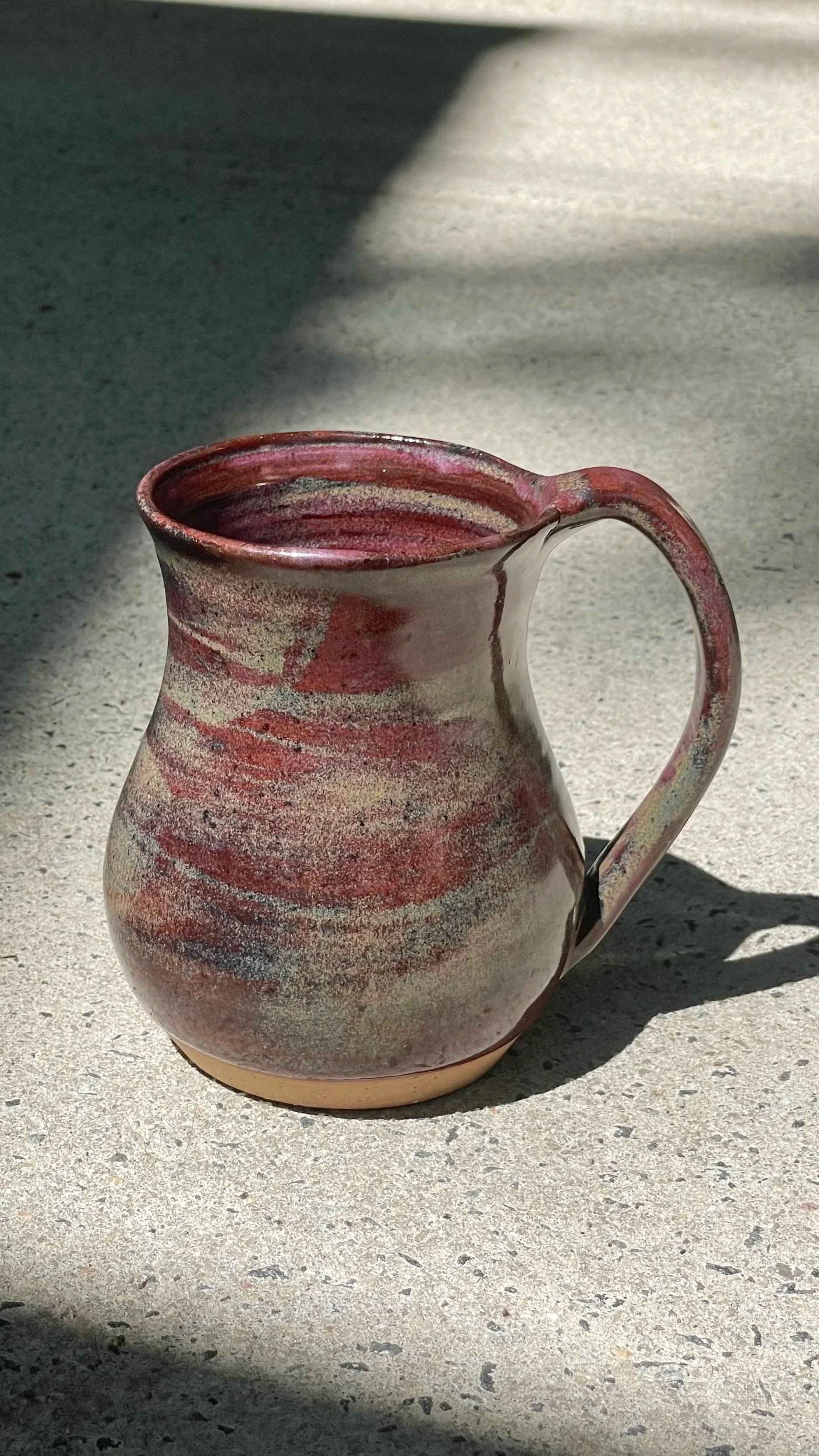 A ceramic mug with a reddish-brown, beige, and gray glaze design, placed on a concrete surface with shadows cast from sunlight.