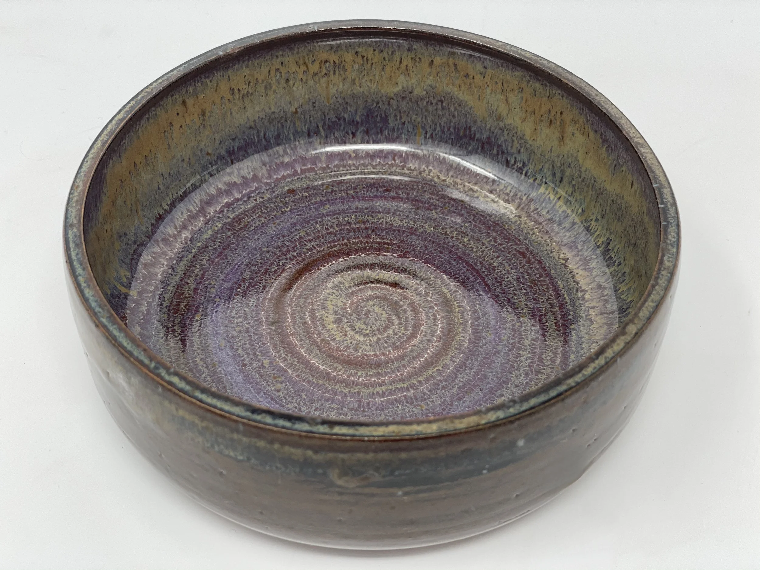A round ceramic bowl with a glazed interior featuring a spiral pattern in purple, brown, and yellow hues, placed on a white surface.