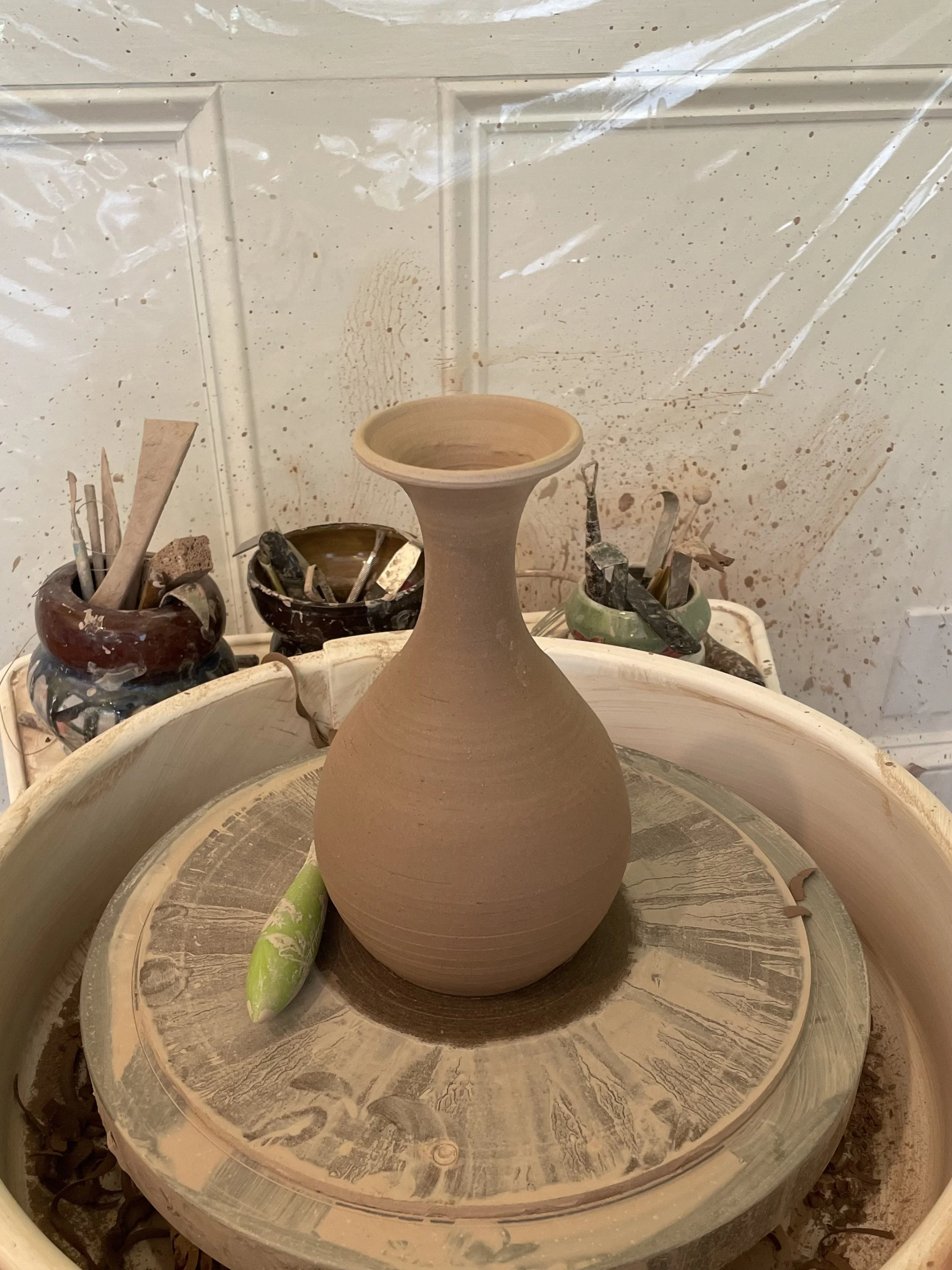 A ceramic vase in progress on a pottery wheel, surrounded by pottery tools and clay splatters in a pottery studio.