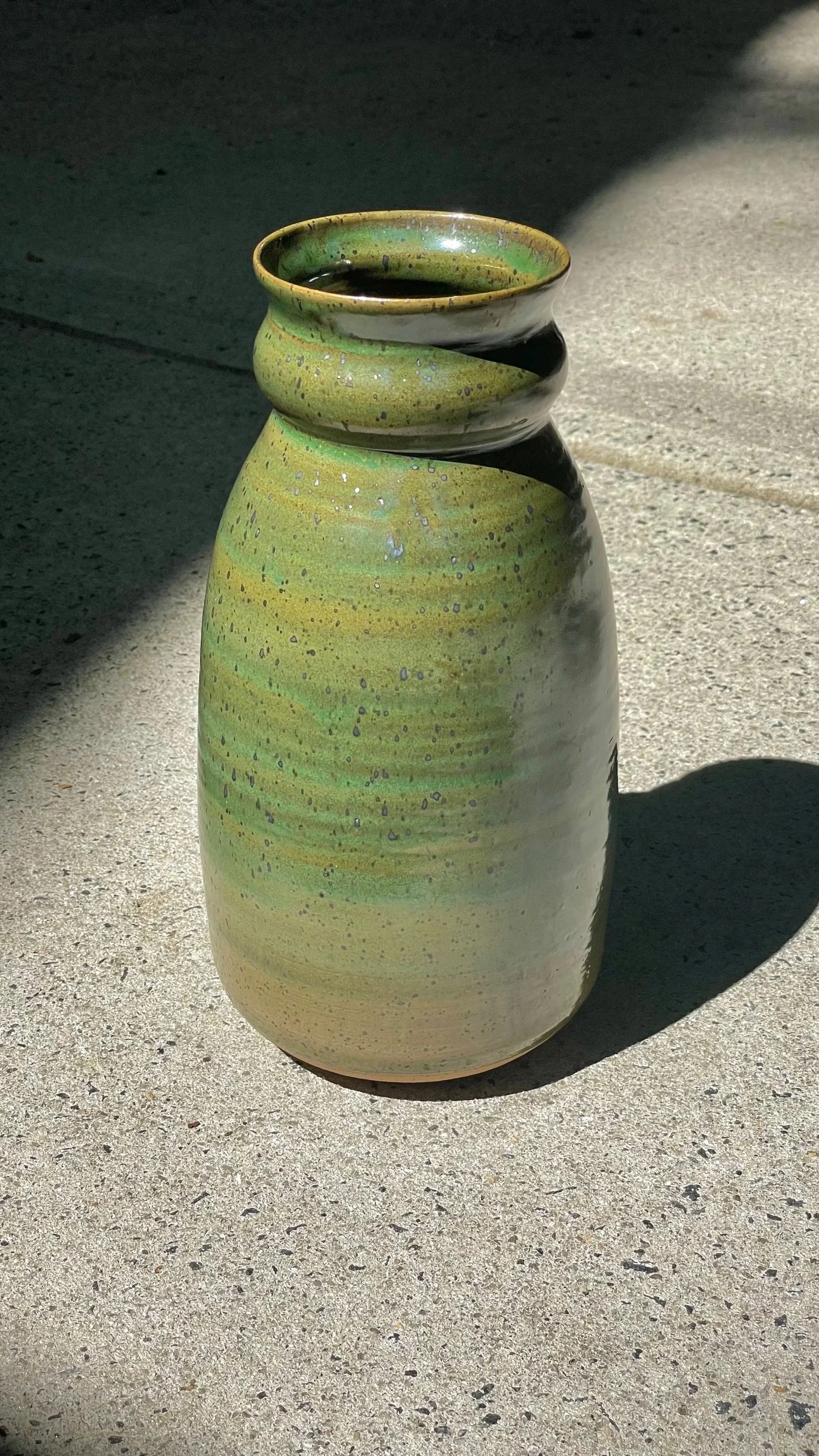 A green ceramic flower vase with a gradient glaze from light beige at the bottom to green at the top, standing on a concrete surface with sunlight and shadows.