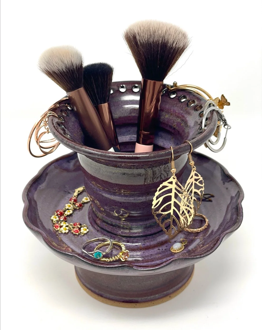 A purple ceramic jewelry holder with makeup brushes and jewelry, including rings and earrings.